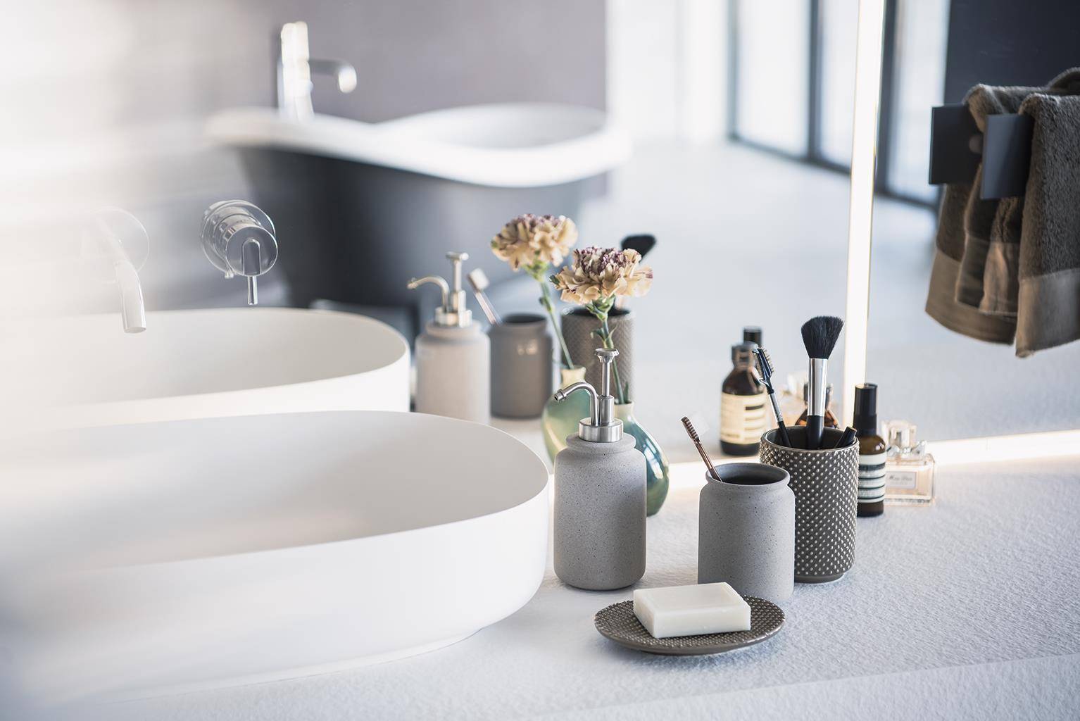 Modernes Badezimmerwaschtisch mit zwei weißen Waschbecken, Seife und Toilettenartikeln auf der Arbeitsplatte, eine Vase mit Blumen und Handtücher, die neben dem Spiegel hängen.
