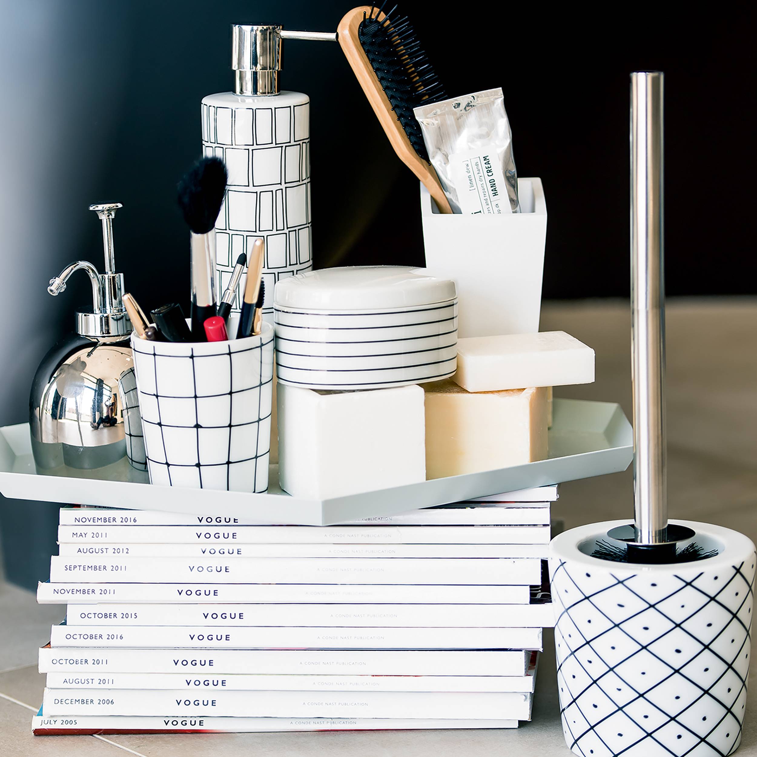 Stilvolles Badezimmer-Waschtisch-Set auf gestapelten Vogue-Magazinen, mit weißen gemusterten Behältern, die Seife, Zahnbürsten, Kosmetika und eine Toilettenbürste enthalten.