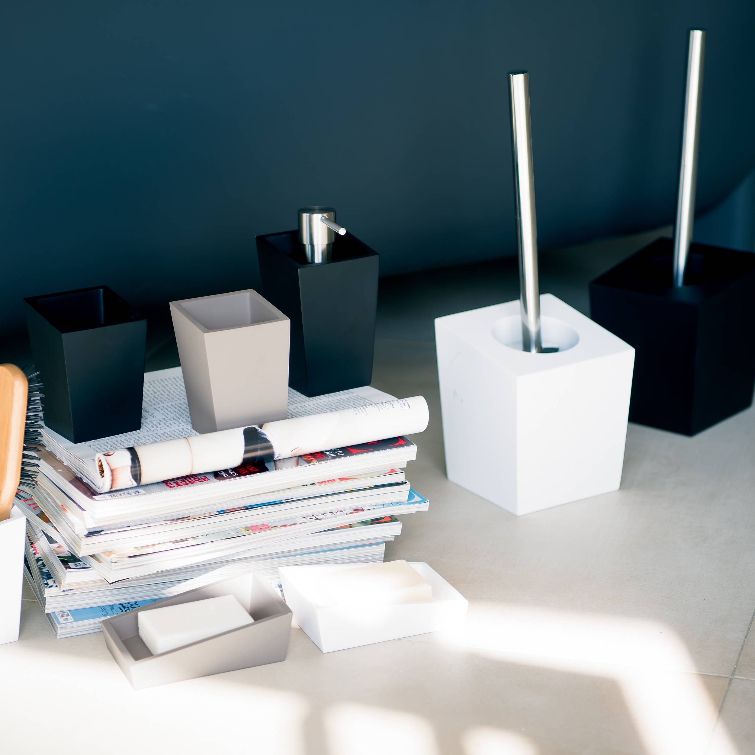 Modernes Büroschreibtisch-Setup mit gestapelten Magazinen, Papierkörben und mehreren quadratischen Stifthaltern in Schwarz, Weiß und Grau, beleuchtet durch helles Sonnenlicht.