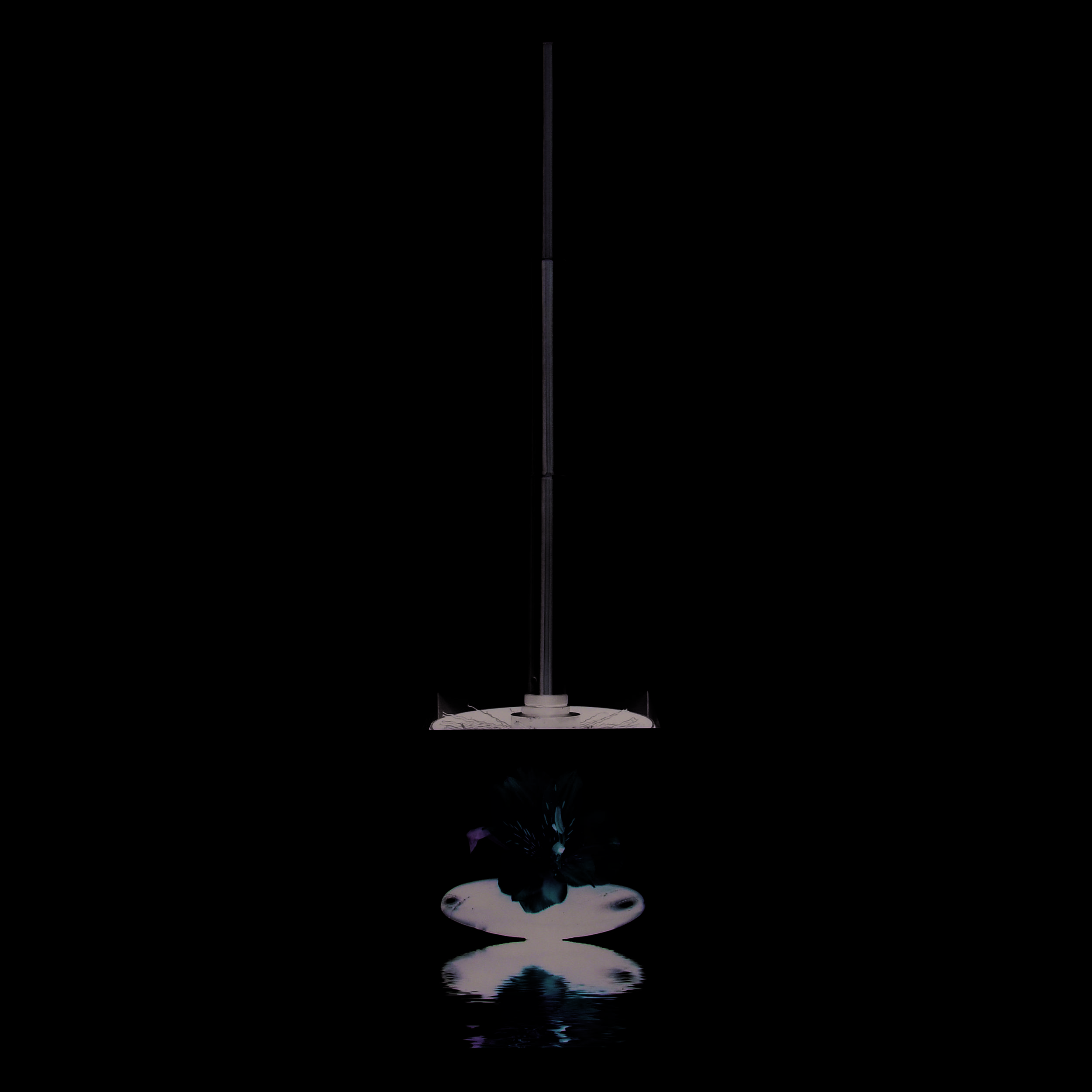 Dekorativer Tischbrunnen mit einem hohen Edelstahlpfosten, der sich von einer weißen würfelförmigen Basis erhebt, mit Blumenmotiv und Wasserreflektion.