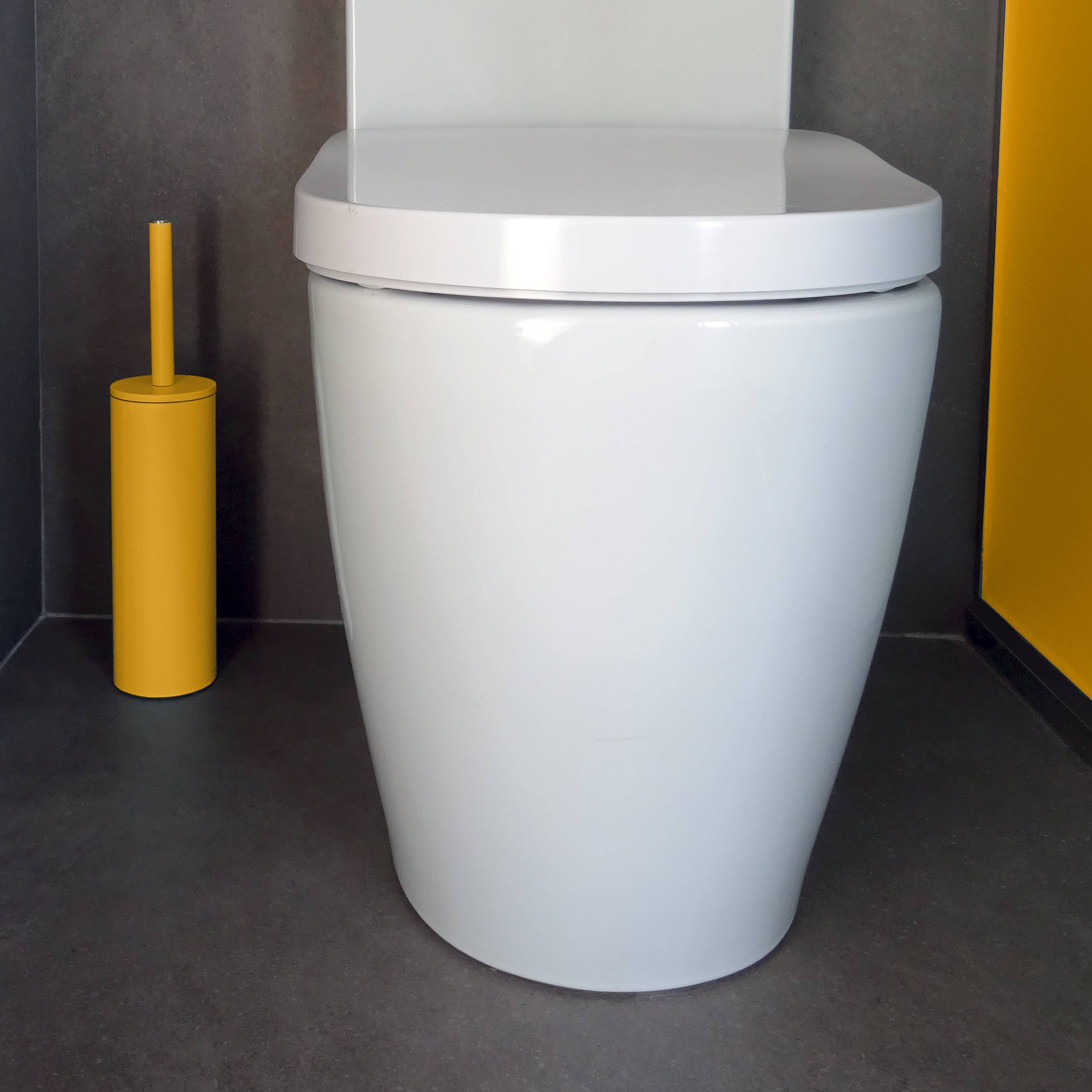 Modernes weißes Toilett mit geschlossenem Deckel in einem grau-gelben Badezimmer, daneben ein gelber Toilettenbürsten-Halter.