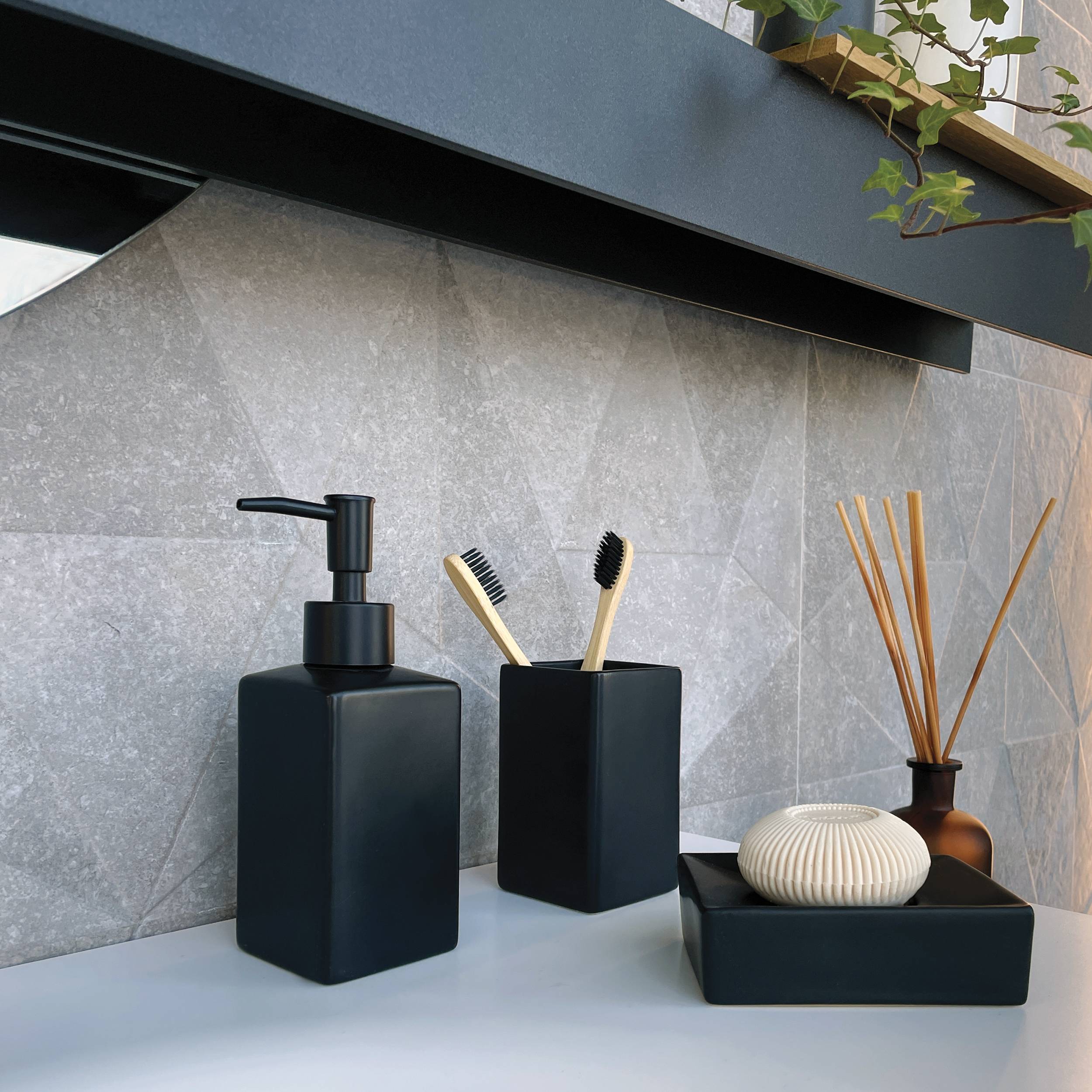 Modernes Badezimmer-Waschbeckenplatte mit schwarzem Seifenspender, Zahnbürstenhalter, Raumduft-Diffusor und kleinem dekorativem Tablett vor einer grauen gekachelten Rückwand.