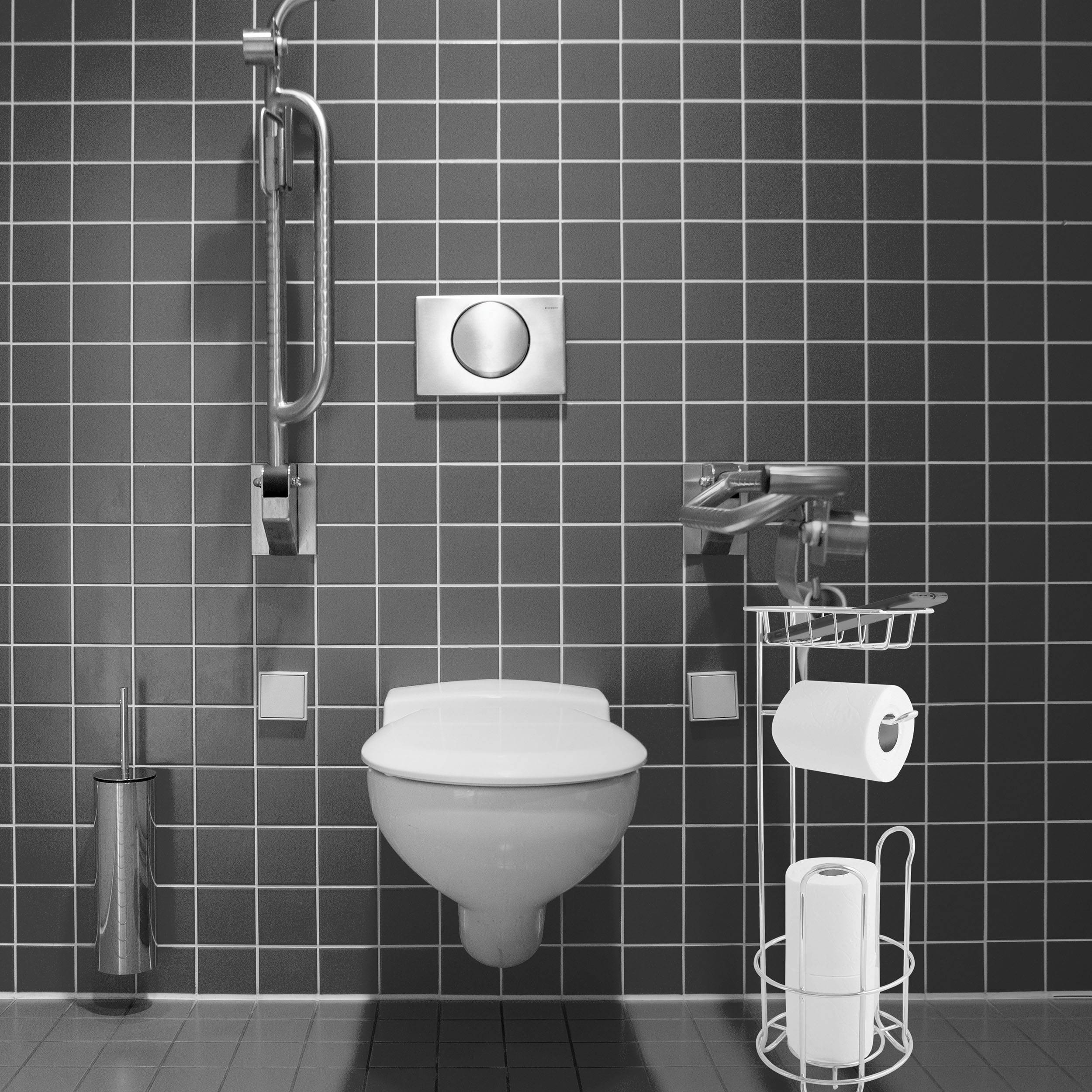 Modernes Badezimmer mit Fliesen, an der Wand montiertem Toilettenbecken, Haltegriff, Spülknopf, Toilettenbürste und Toilettenpapierhalter.