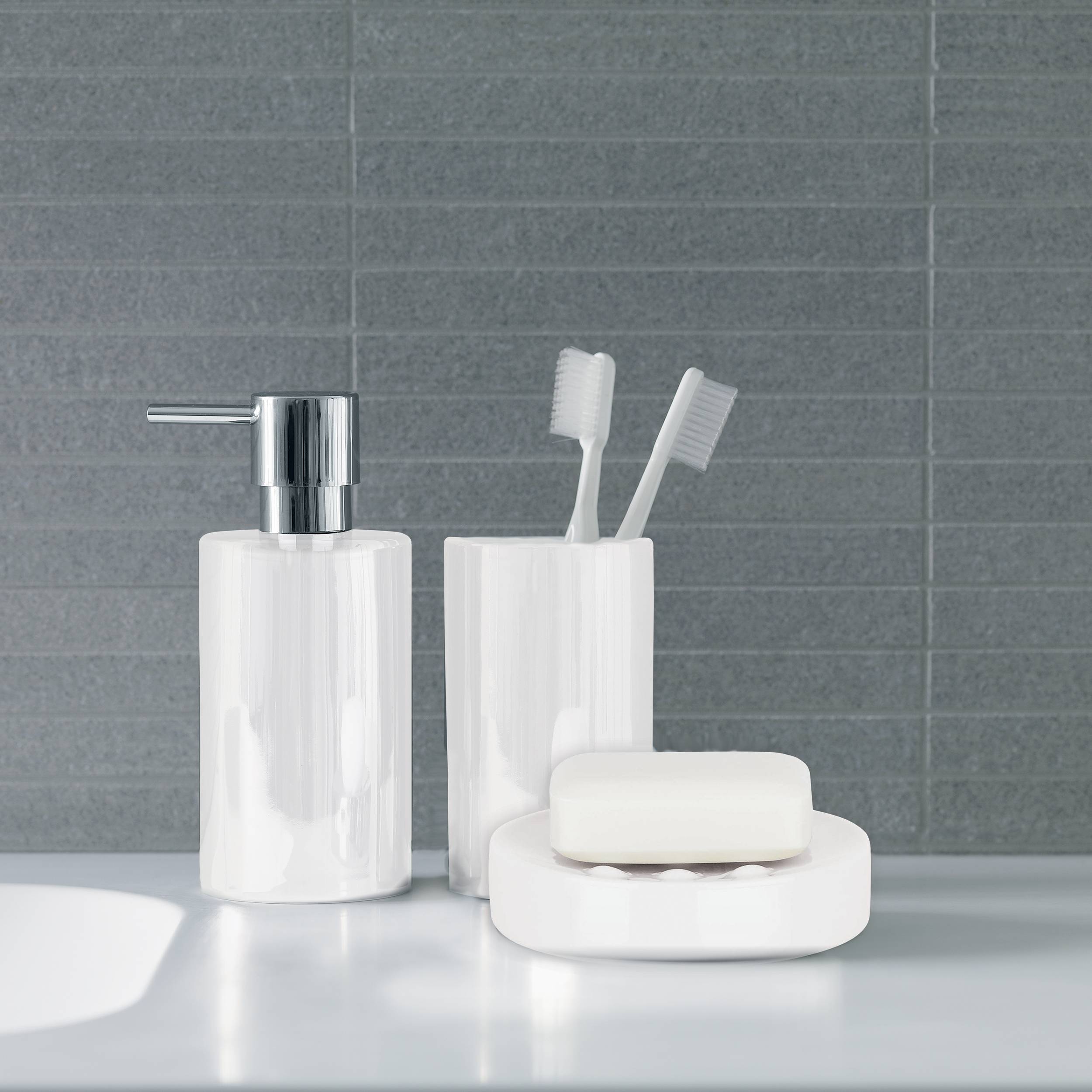 Badezimmerablage mit weißem Seifenspender, Zahnbürstenhalter mit zwei Zahnbürsten und Seifenschale vor grauer gekachelter Wand.