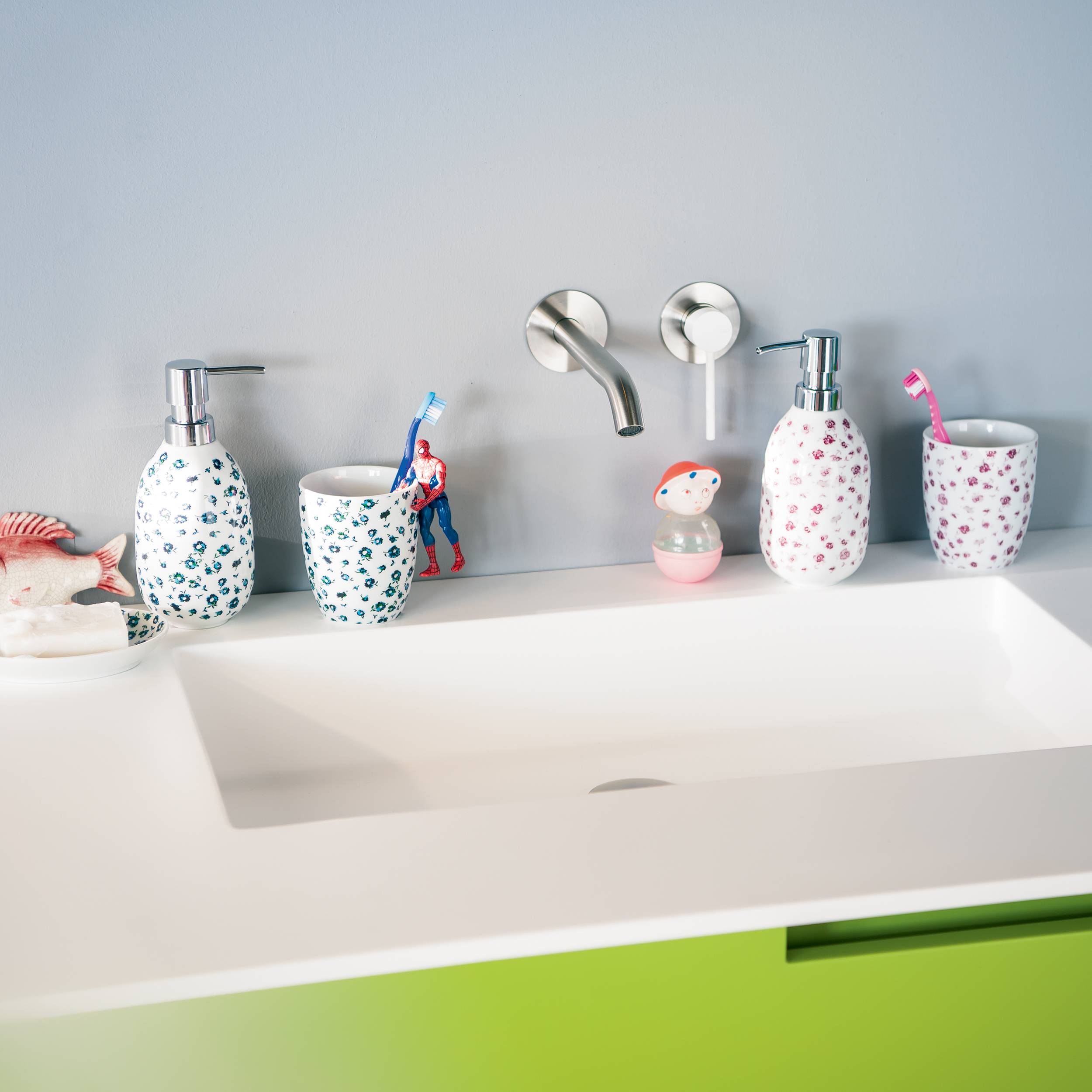 Modernes Badezimmerbecken mit zwei wandmontierten Wasserhähnen und passenden gemusterten Seifenspendern, Bechern und Zahnbürstenhaltern auf einer weißen Arbeitsplatte über einem grünen Schrank.