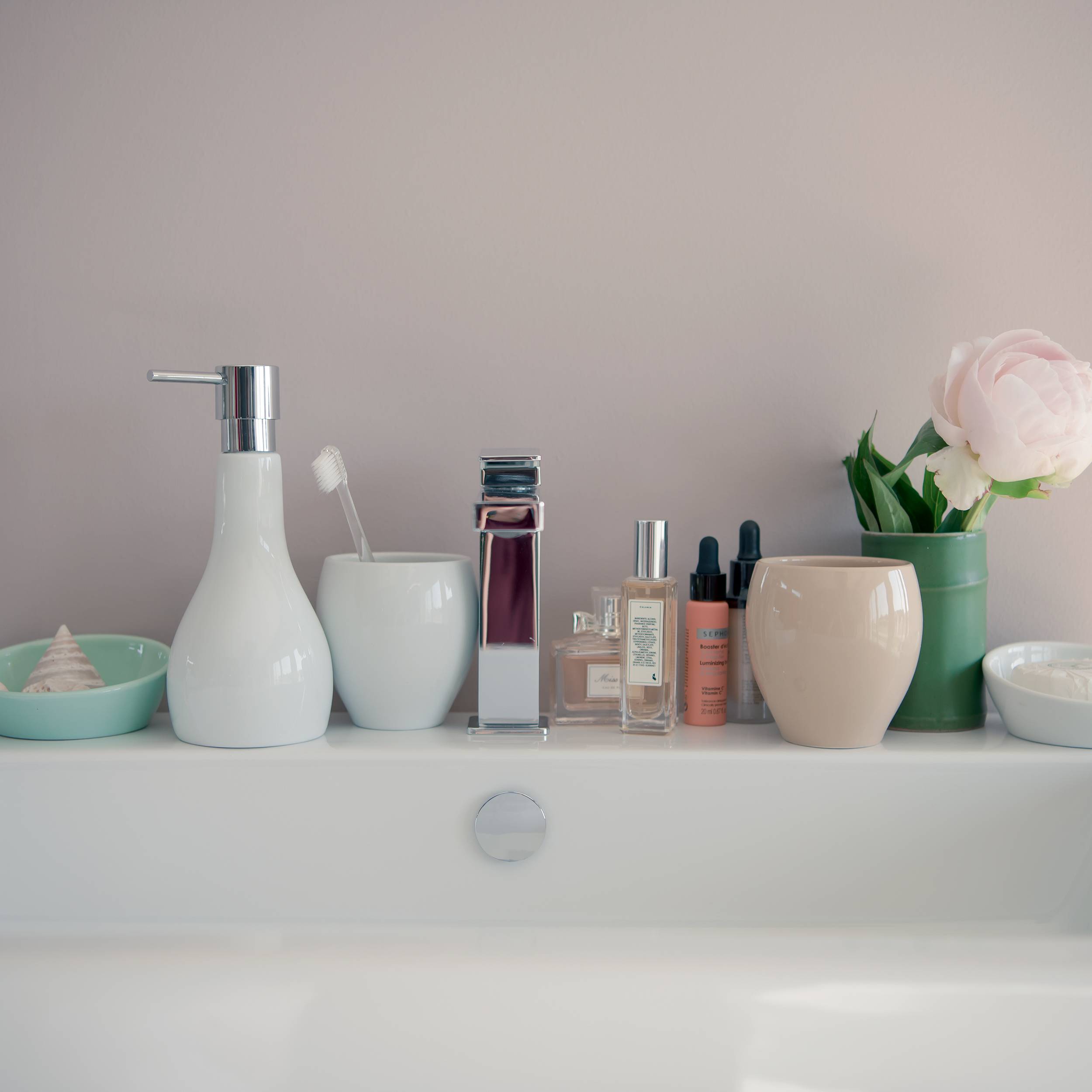 Badezimmer-Waschtischablage mit verschiedenen Toilettenartikeln, Parfümflaschen und Vasen, einschließlich einer rosa Blume in einer grünen Vase und einem Wattestäbchenhalter.
