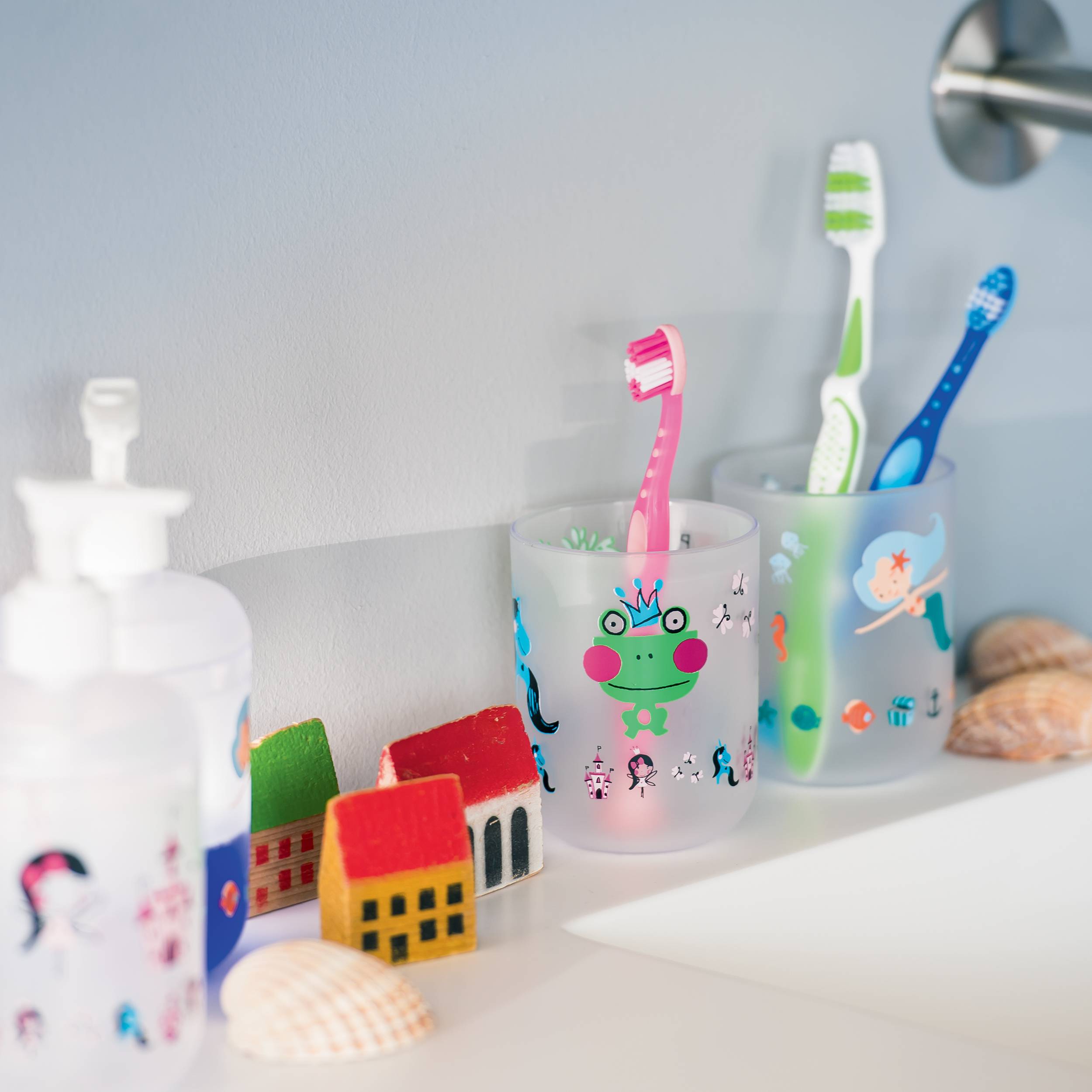 Badezimmerregal mit Zahnbürsten für Kinder in dekorierten Bechern neben einem Seifenspender und kleinen Spielzeughäusern und Muscheln.