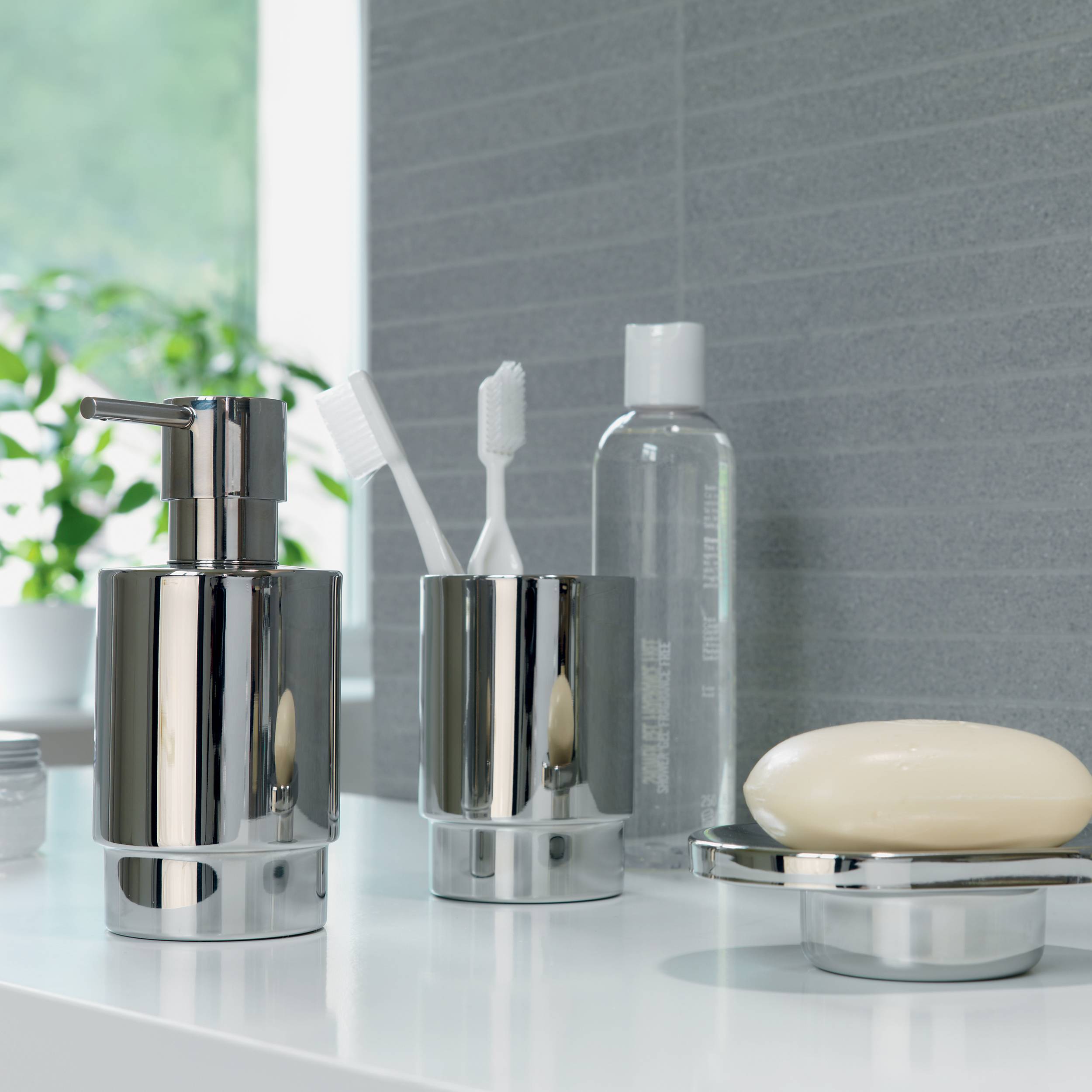 Badezimmer-Arbeitsplatte mit chromglänzendem Seifenspender, Zahnbürstenhalter, klarer Flasche und Seifenstück auf einem Tablett.