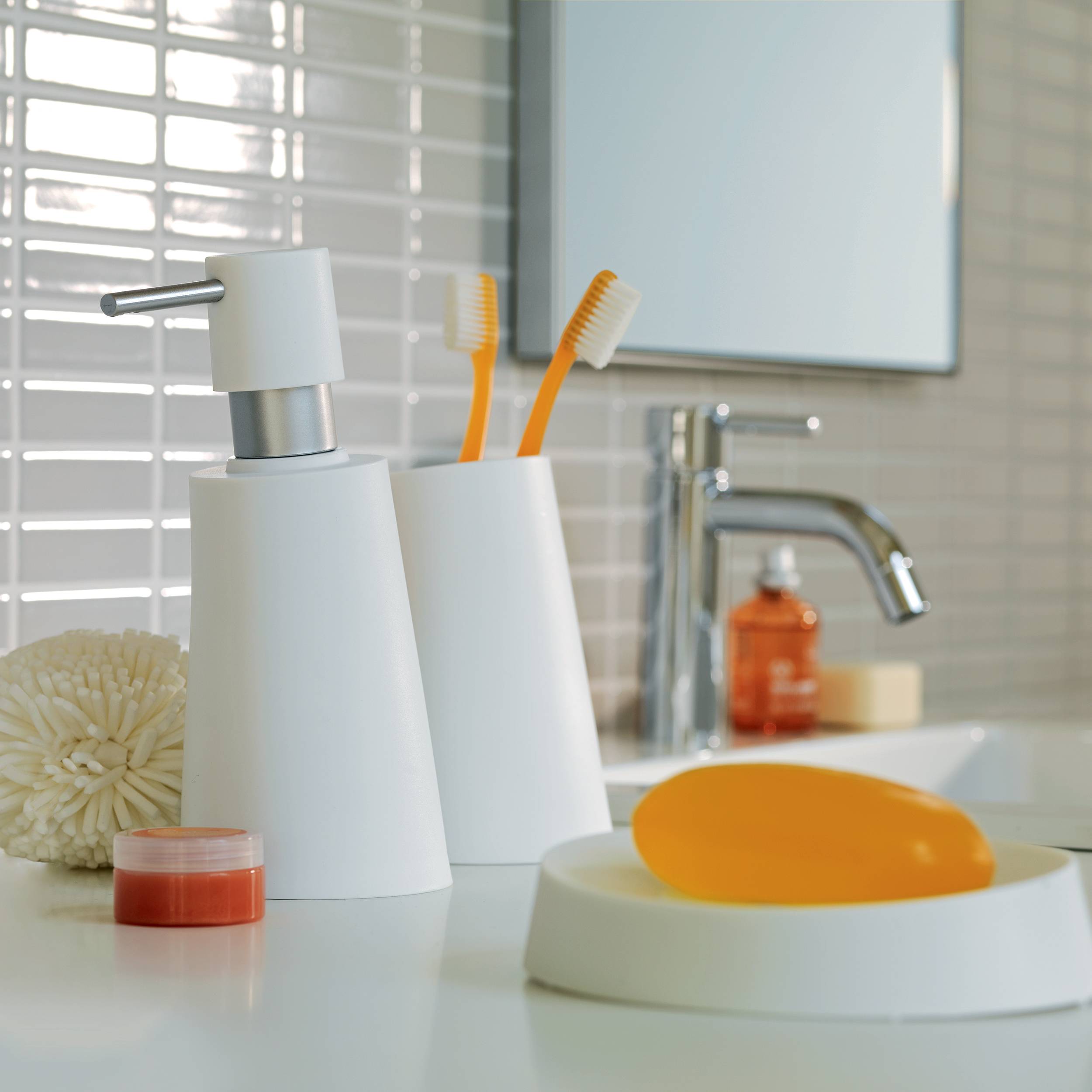 Badezimmerwaschbeckenablage mit weißen Seifenspendern, Zahnbürsten und einem Stück orangefarbener Seife neben einem Chromwasserhahn.