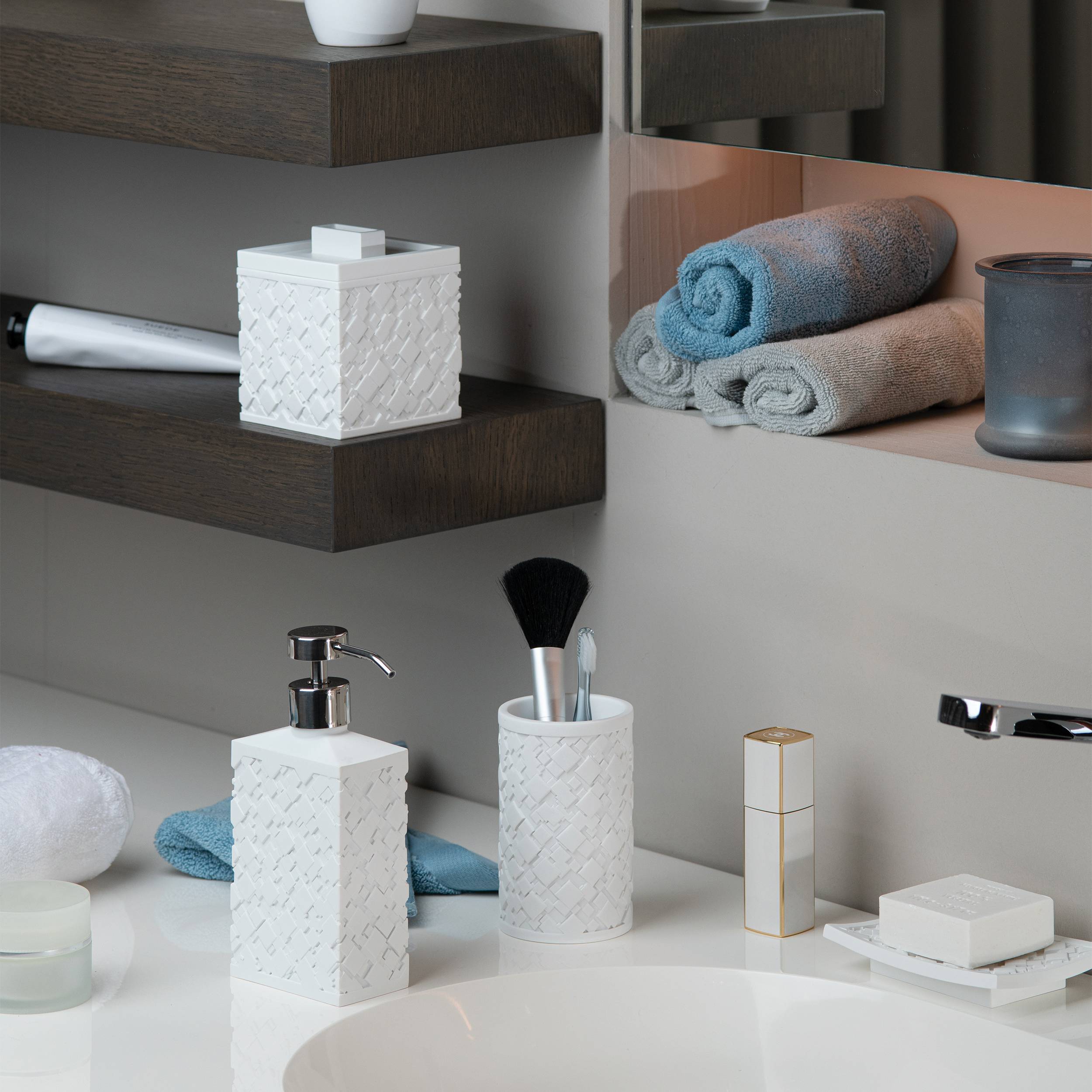 Eine ordentlich angeordnete Badezimmerablage mit weißen strukturierten Behältern, Seifenspender, Makeup-Pinseln, gefalteten Handtüchern auf Regalen und einem Waschbecken im Vordergrund.