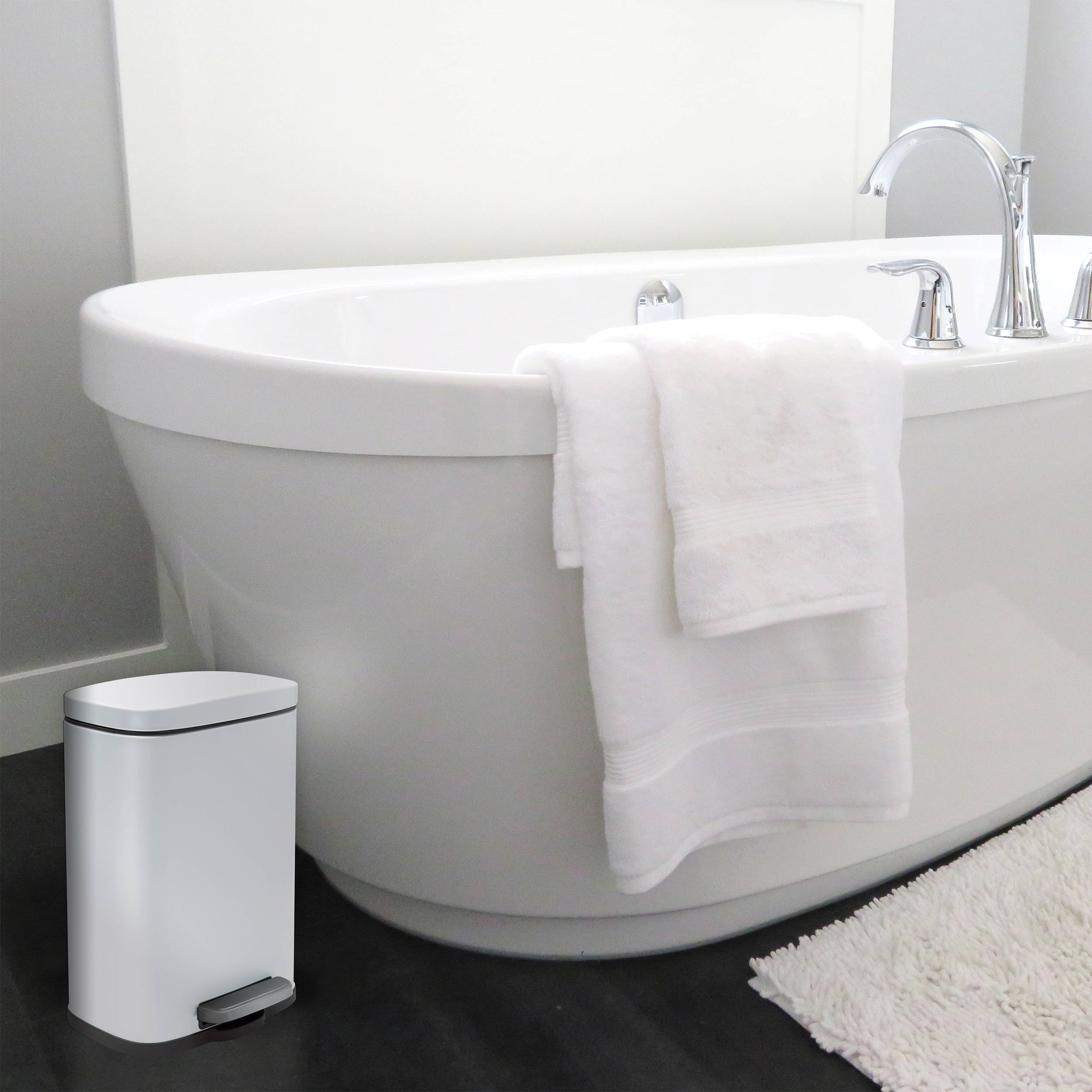 Freistehende weiße Badewanne mit Chromarmatur und ein weißes Handtuch über der Kante drapiert, neben einem kleinen weißen Badezimmereimer auf dunklem Boden.