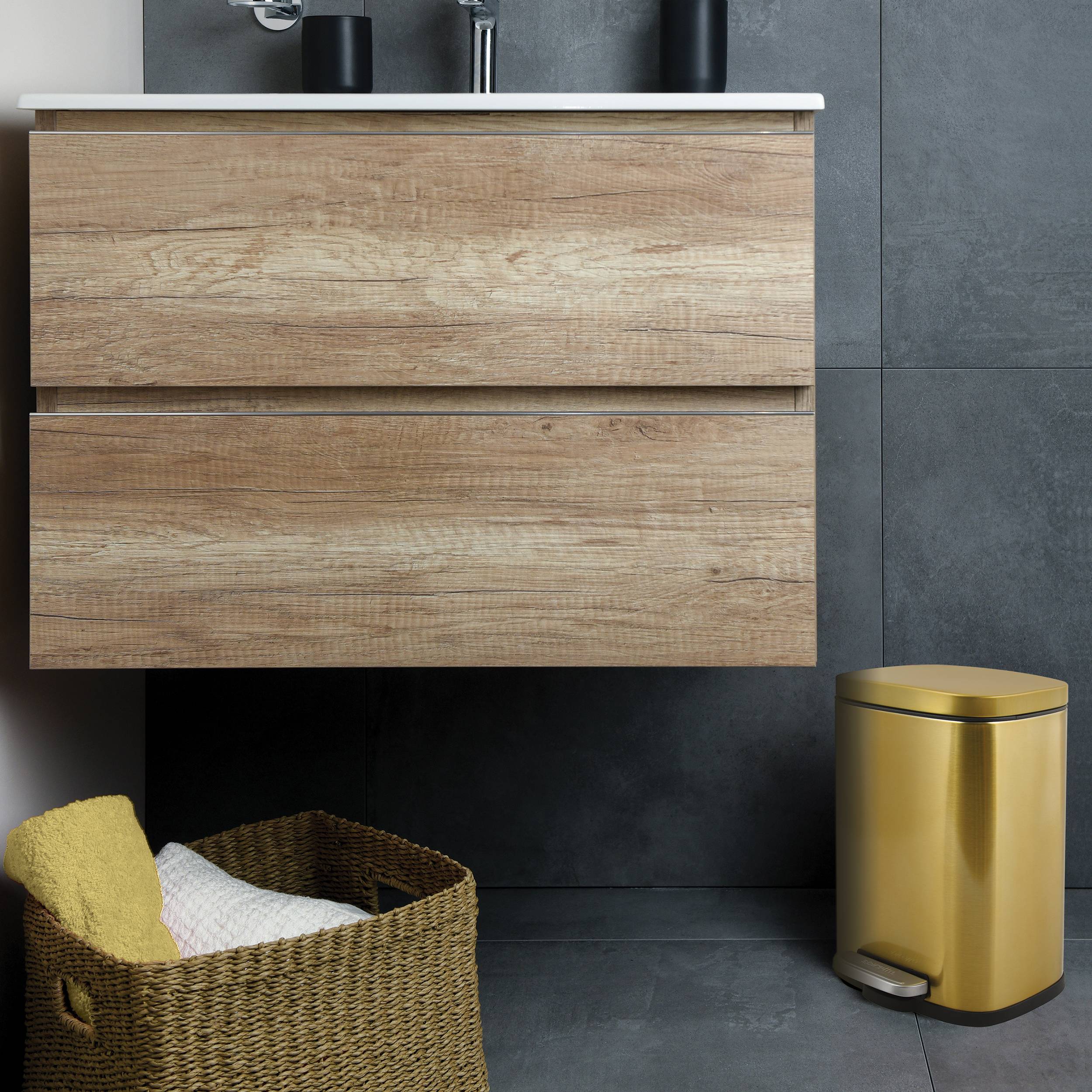 Modernes Badezimmerwaschtisch mit zwei Holzschubladen, geflochtenem Wäschekorb und goldenem Papierkorb neben einer dunkel gefliesten Wand.