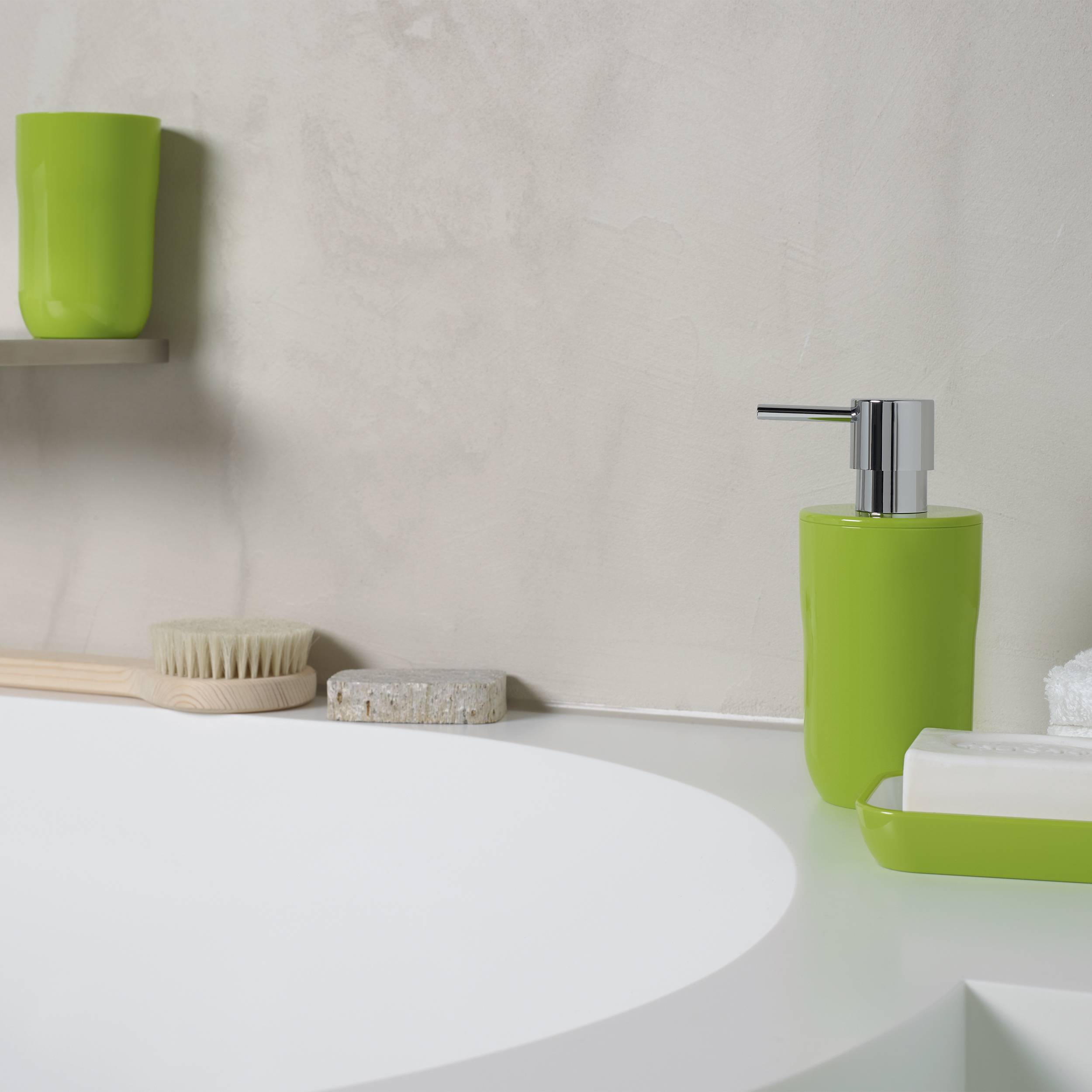 Minimalistisches Badezimmerbecken mit leuchtend grünem Seifenspender, Becher, Tablett und Bürste auf einer weißen Arbeitsplatte vor einer schlichten Wand.