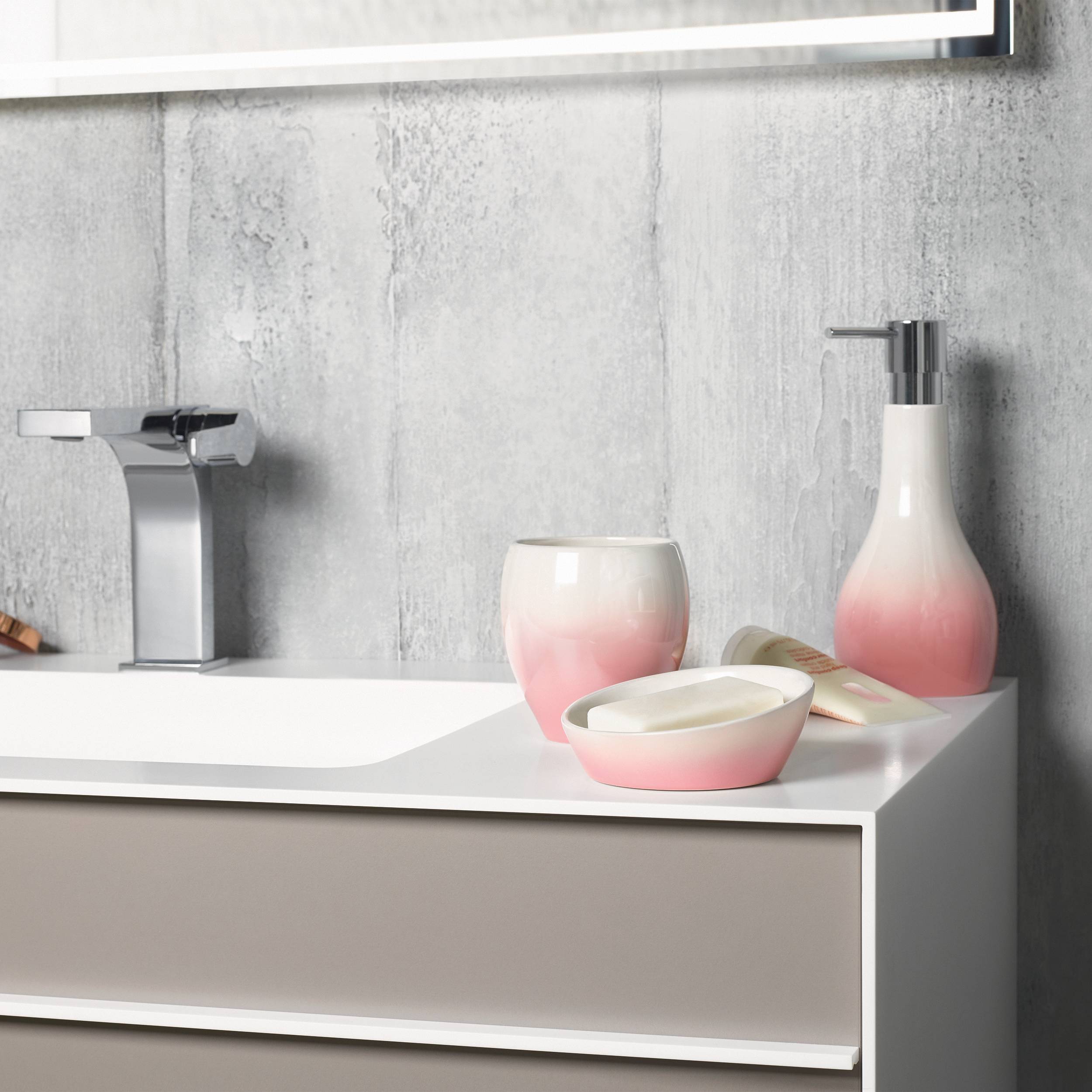 Modernes Badezimmerwaschtisch mit verchromtem Wasserhahn, rosa-weißem Seifenspender, Becher und Schale auf einer weißen Arbeitsplatte vor einer grau strukturierten Wand.