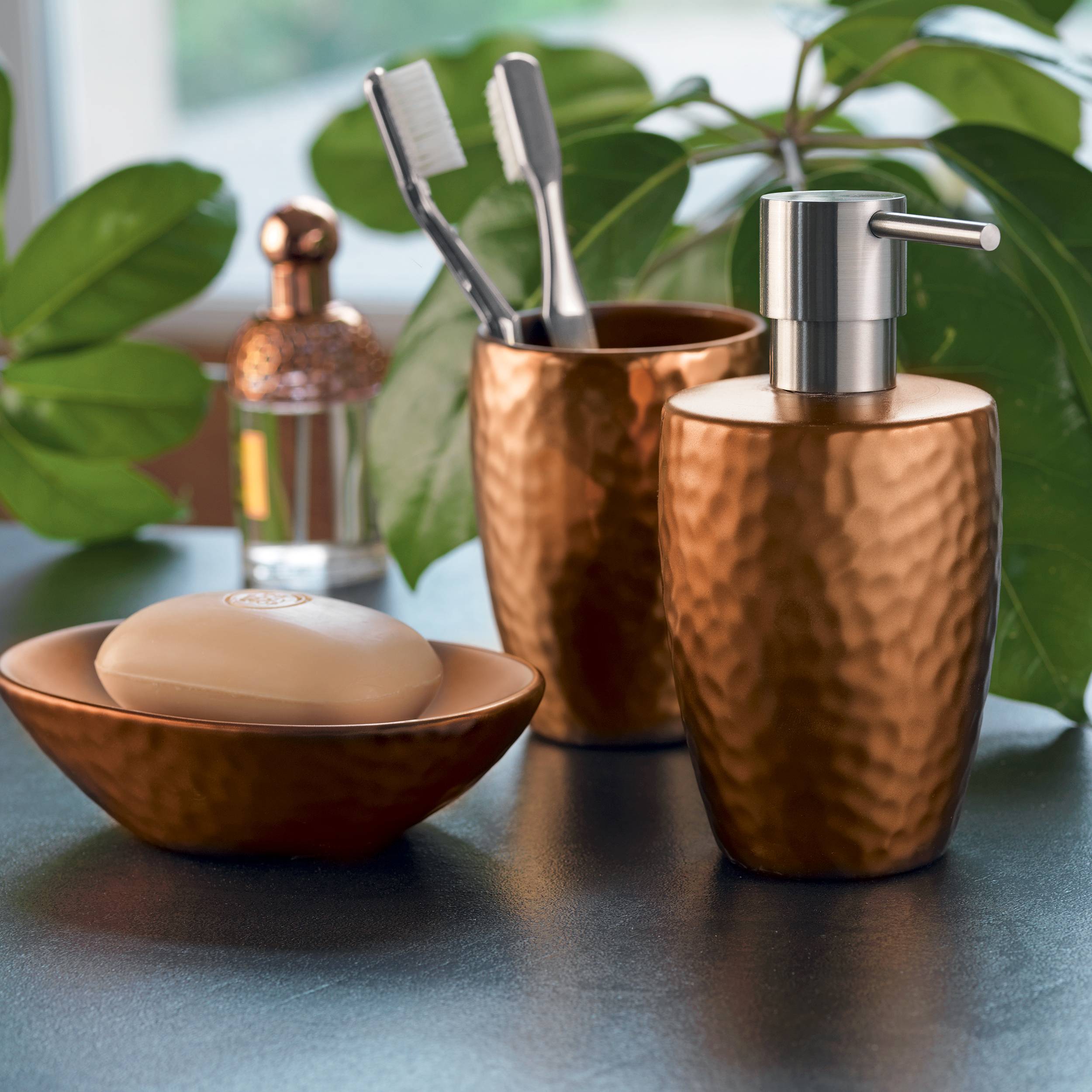 Kupfernes Badezimmer-Accessoires-Set auf einer Arbeitsplatte, bestehend aus einem Seifenspender, Zahnbürstenhalter, Seifenschale und einer kleinen Flasche, mit grünen Pflanzen im Hintergrund.