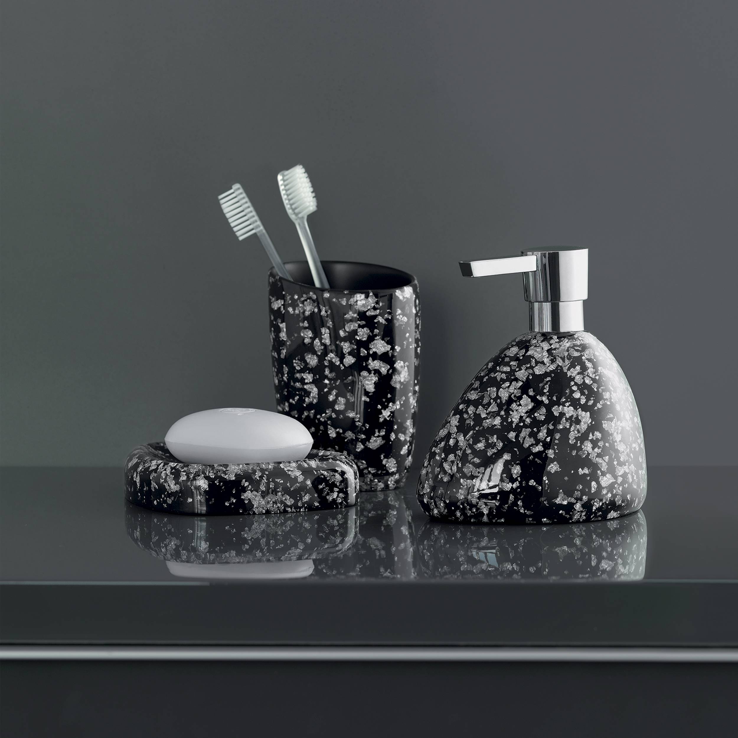 Schwarzes Badezimmer-Accessoire-Set mit Sprenkeldesign bestehend aus einem Seifenspender, einem Zahnbürstenhalter und einer Seifenschale auf einer reflektierenden Oberfläche.