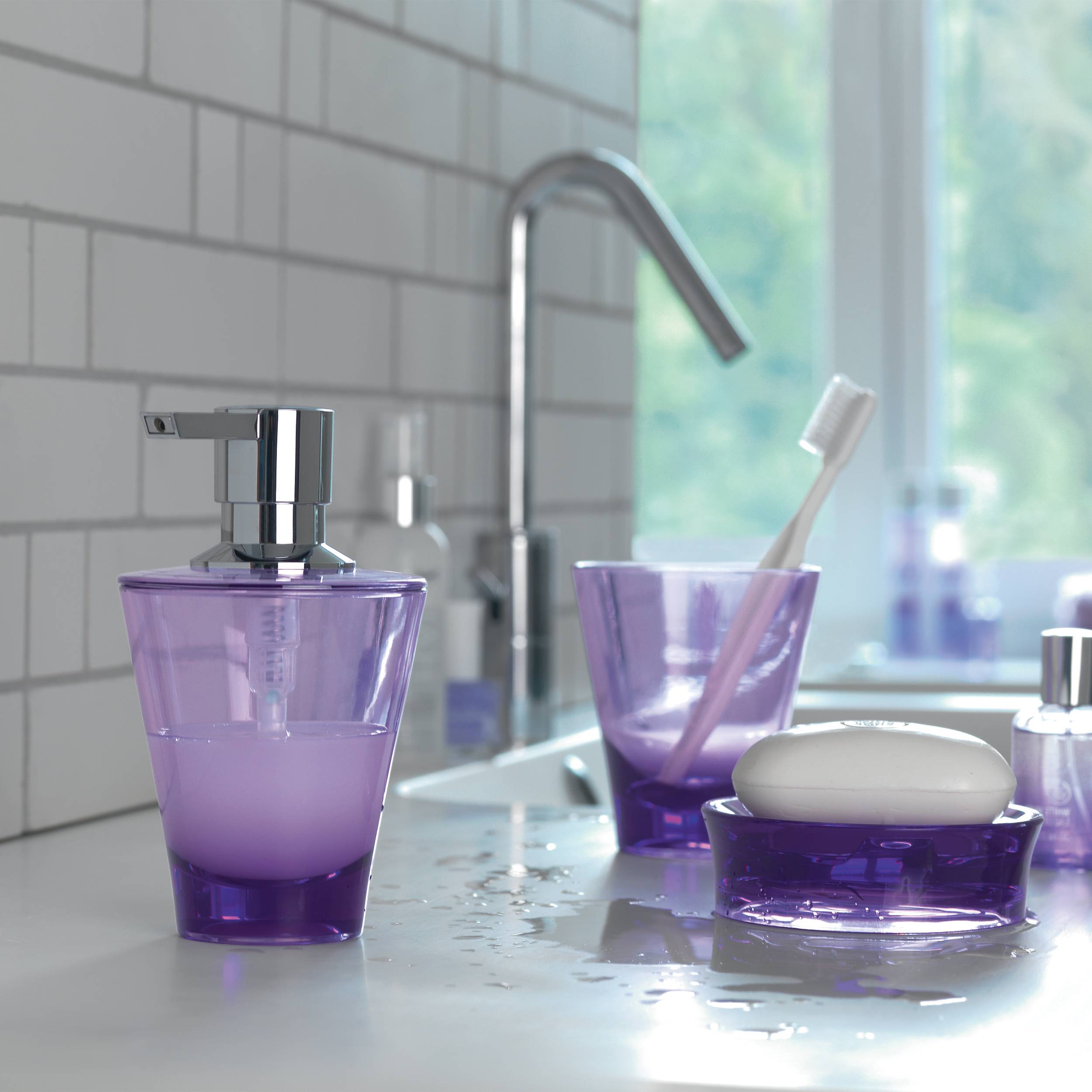 Lila Badezimmerset mit Seifenspender, Zahnbürstenhalter und Seifenschale auf einer nassen Spülenplatte neben einem Wasserhahn und Fenster.