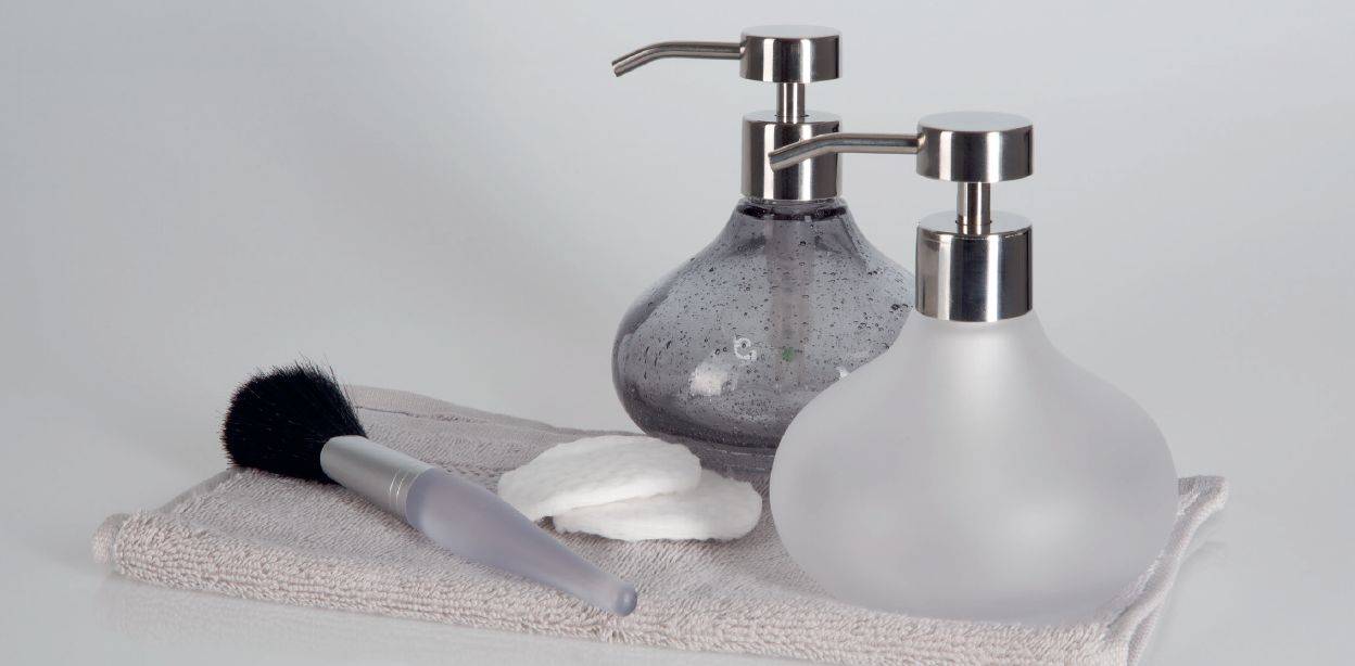 Zwei Pumpflaschen, ein Makeup-Pinsel und ein gefaltetes Handtuch auf einer hellgrauen Oberfläche, was auf ein Badezimmer oder eine Hautpflege-Umgebung hindeutet.