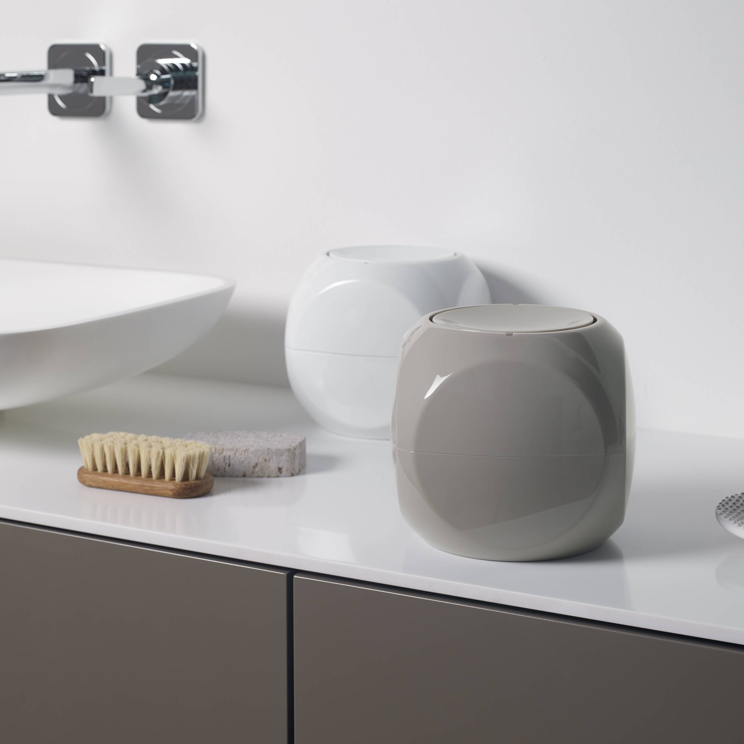 Minimales Badezimmer-Waschtisch mit Waschbecken, Chrom-Armatur, zwei Keramikbehältern und einer Bürste sowie einem Schwamm auf einer weißen Arbeitsplatte.