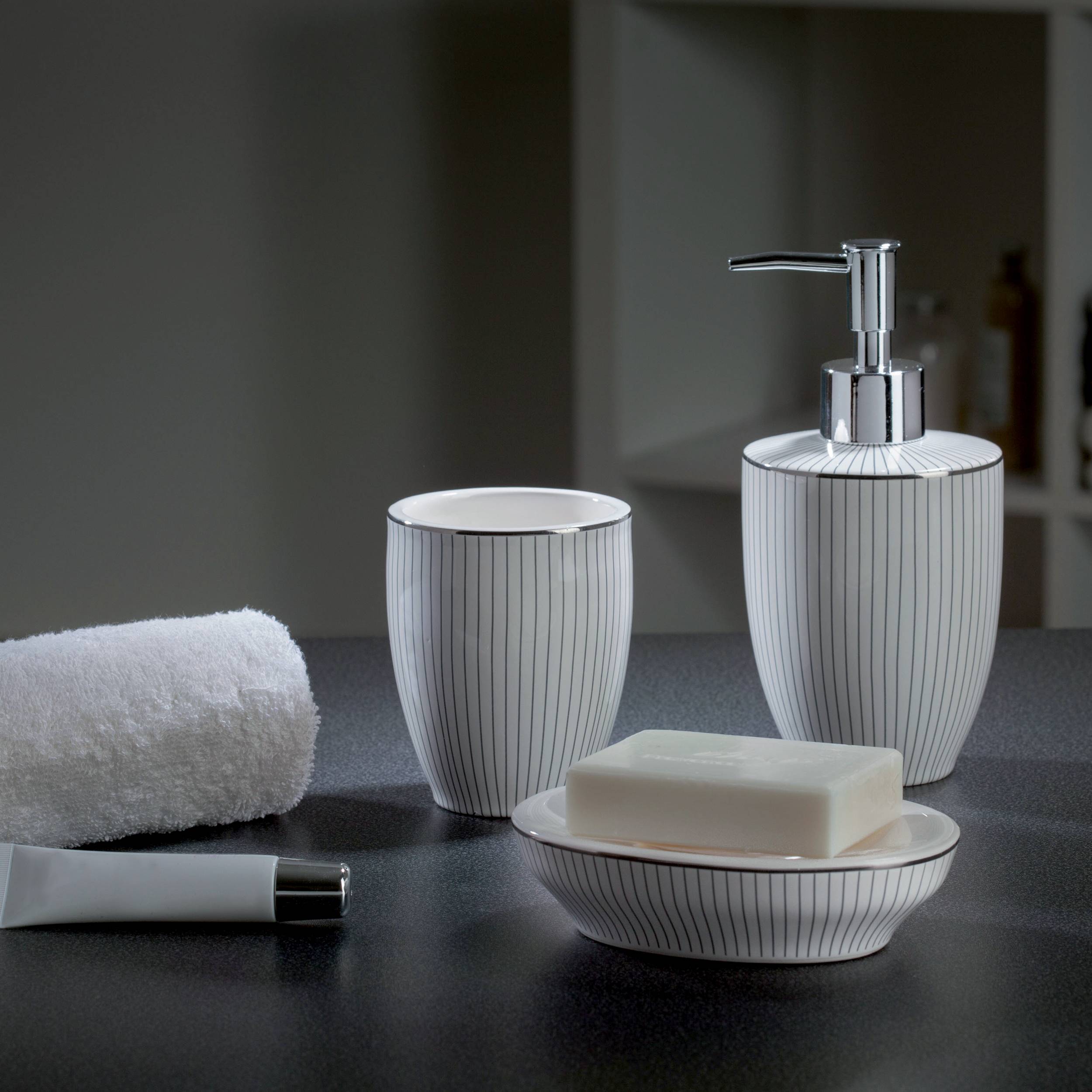 Badezimmer-Arbeitsplattensatz mit gestreiftem Seifenspender, Becher, Schale, Seifenstück, Tube und aufgerolltem Handtuch auf einer dunklen Oberfläche.