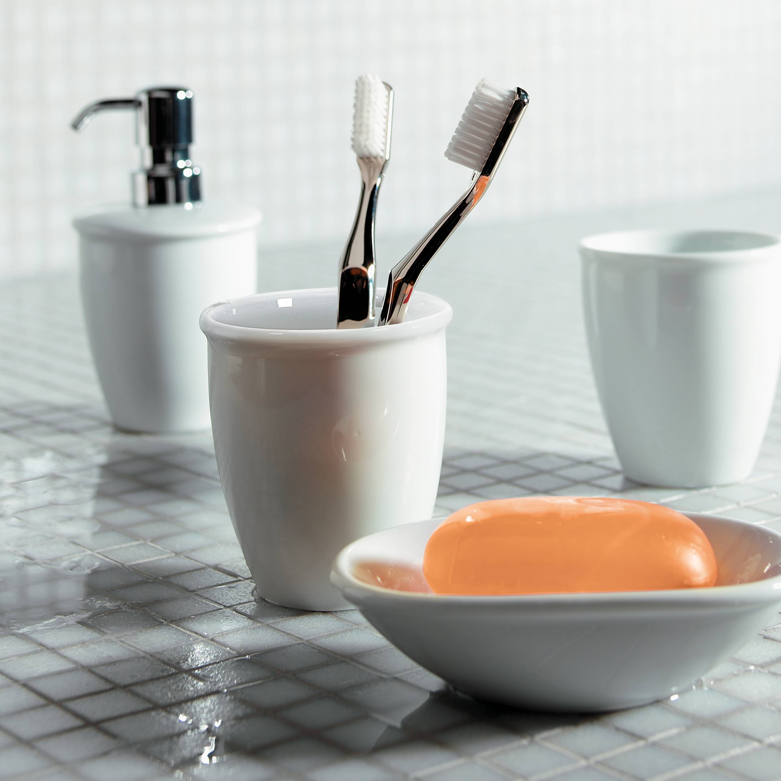Zwei Zahnbürsten in einem weißen Becher neben einem Seifenspender, leeren Becher und einer orangefarbenen Seife auf einer Badezimmerablage.