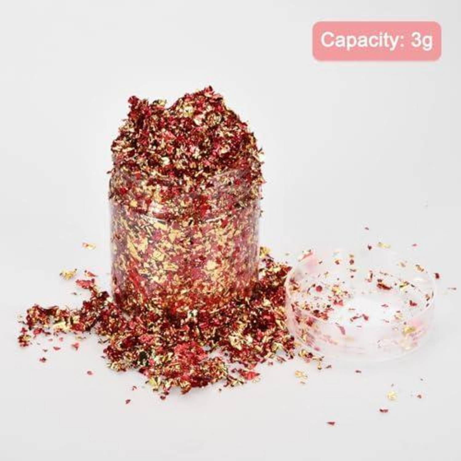 Ein kleines durchsichtiges Glas gefüllt mit rot und goldenem Glitter, mit verschütteltem Glitter rundherum, beschriftet mit „Kapazität: 3g".