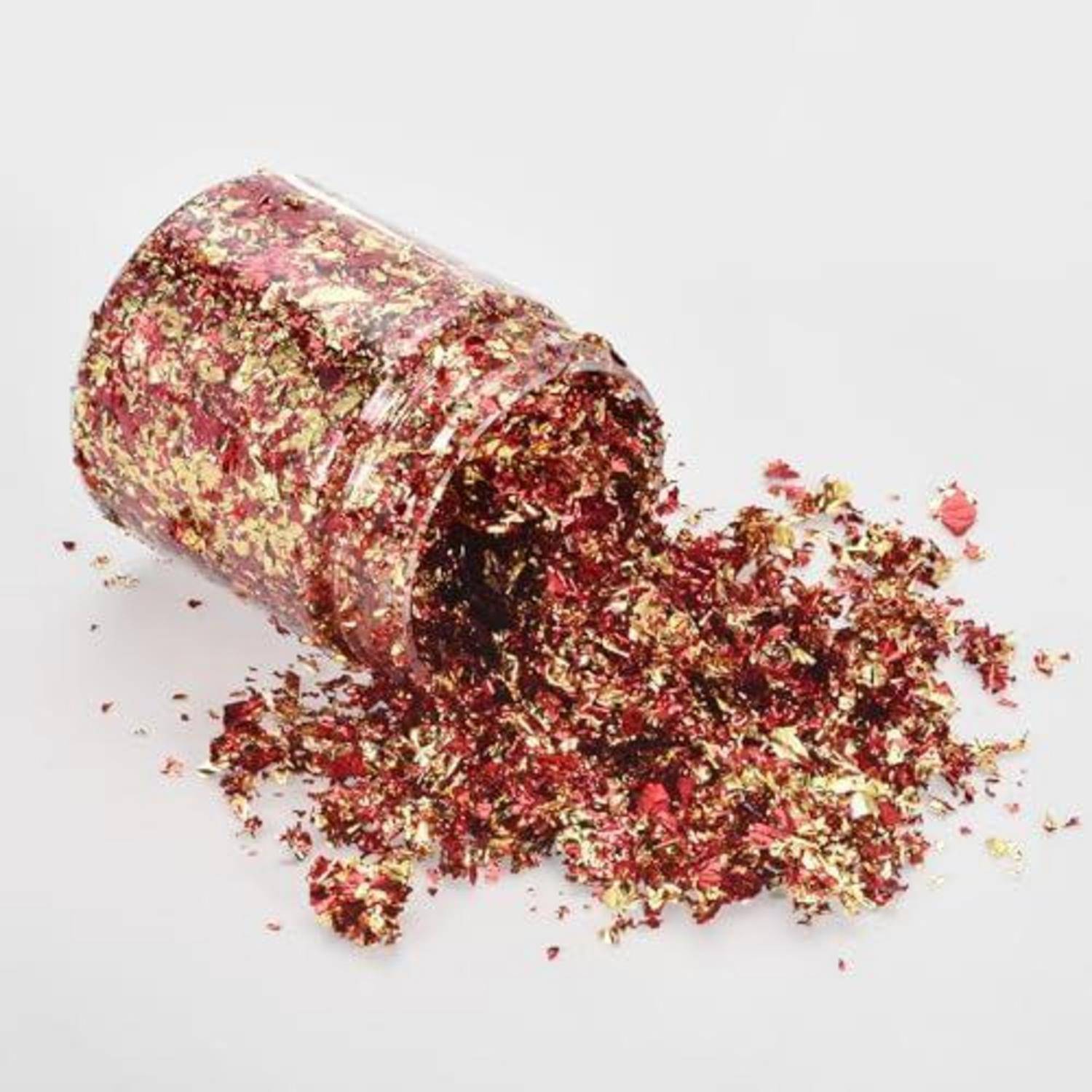 Ein umgestürzter, mit Glitter bedeckter Becher, aus dem rote und goldene Konfetti auf eine weiße Oberfläche verschüttet werden.