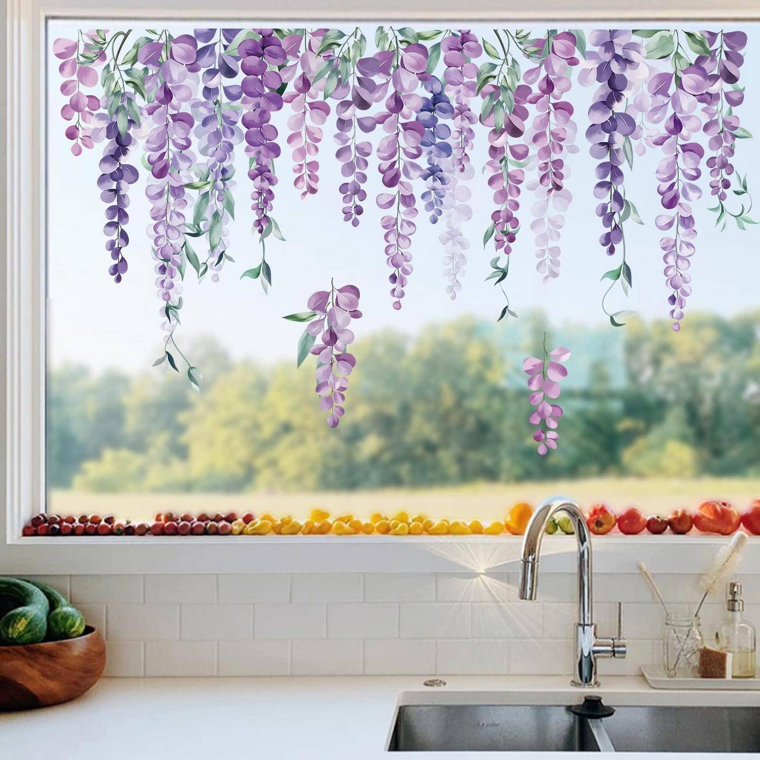 Küchenfenster mit hängenden Blumengirlanden in Violett und Rosa über einem Waschbecken, mit Obst entlang der Fensterbank aufgereiht und Gartenblick nach außen.