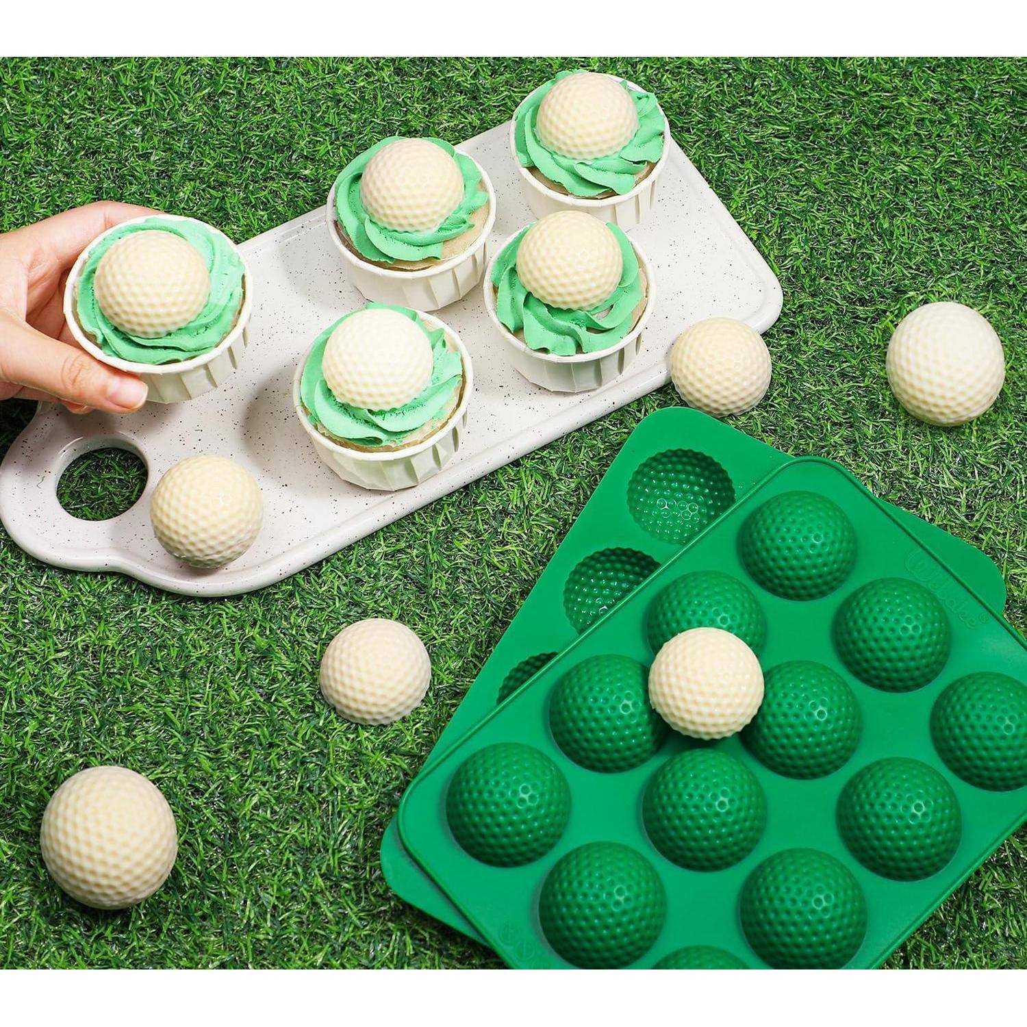 Hand, die golfballförmige Leckereien in Silikonformen auf Gras platziert, mit zusätzlichen Bällen und grünen und weißen Tabletts in der Nähe.
