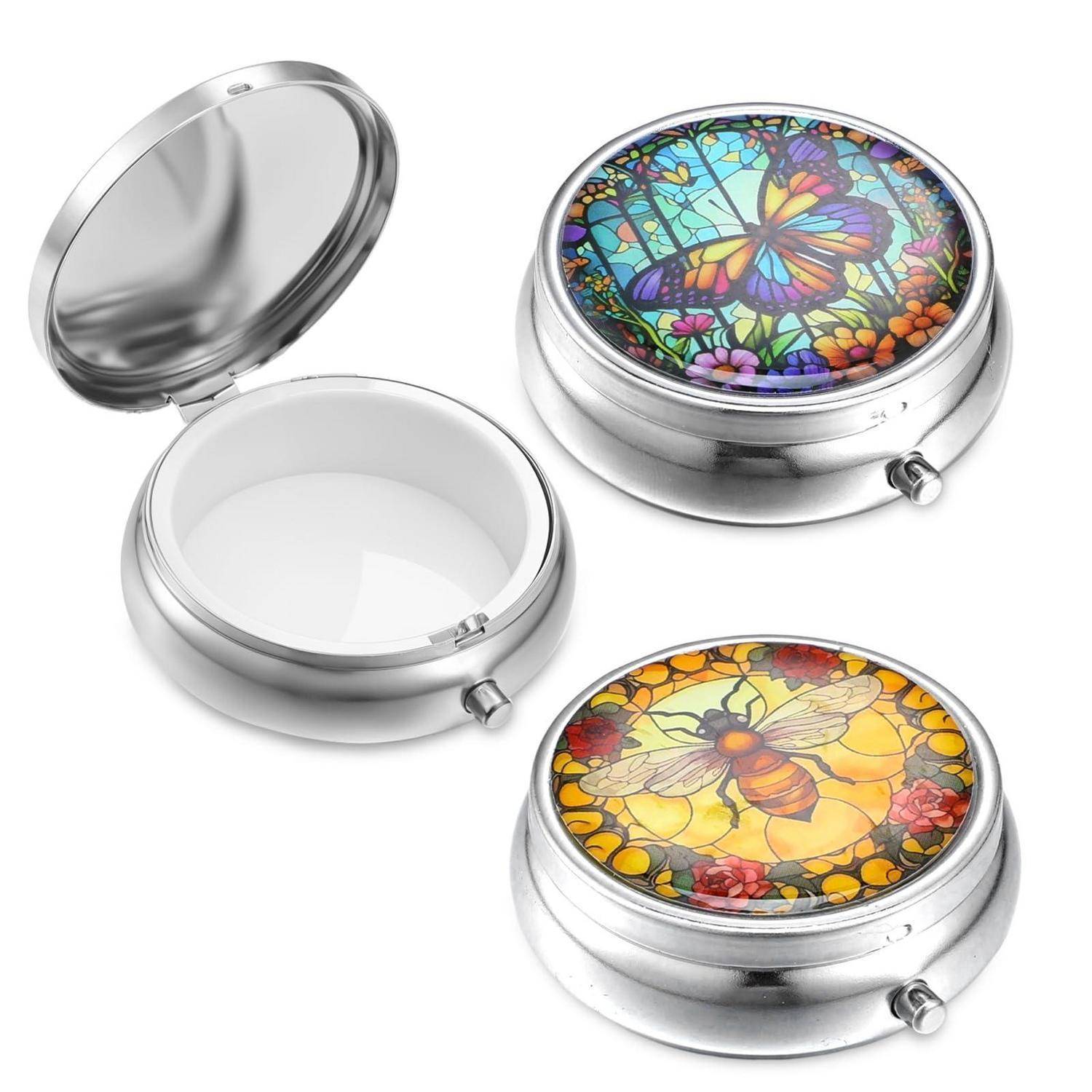 Drei runde Metallpillendosen, eine offene und leer und zwei geschlossene mit farbenfrohen Schmetterlings- und Bienenglasmalerei-Designs.