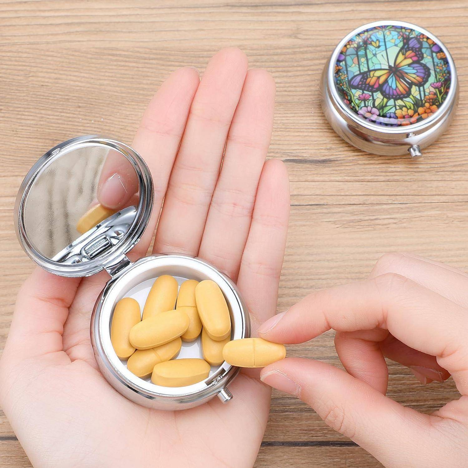 Eine Hand hält ein kleines, rundes Pillendöschen mit gelben Kapseln, während eine andere Hand eine herausnimmt; der Deckel des Döschen mit Schmetterlingsdesign liegt in der Nähe.