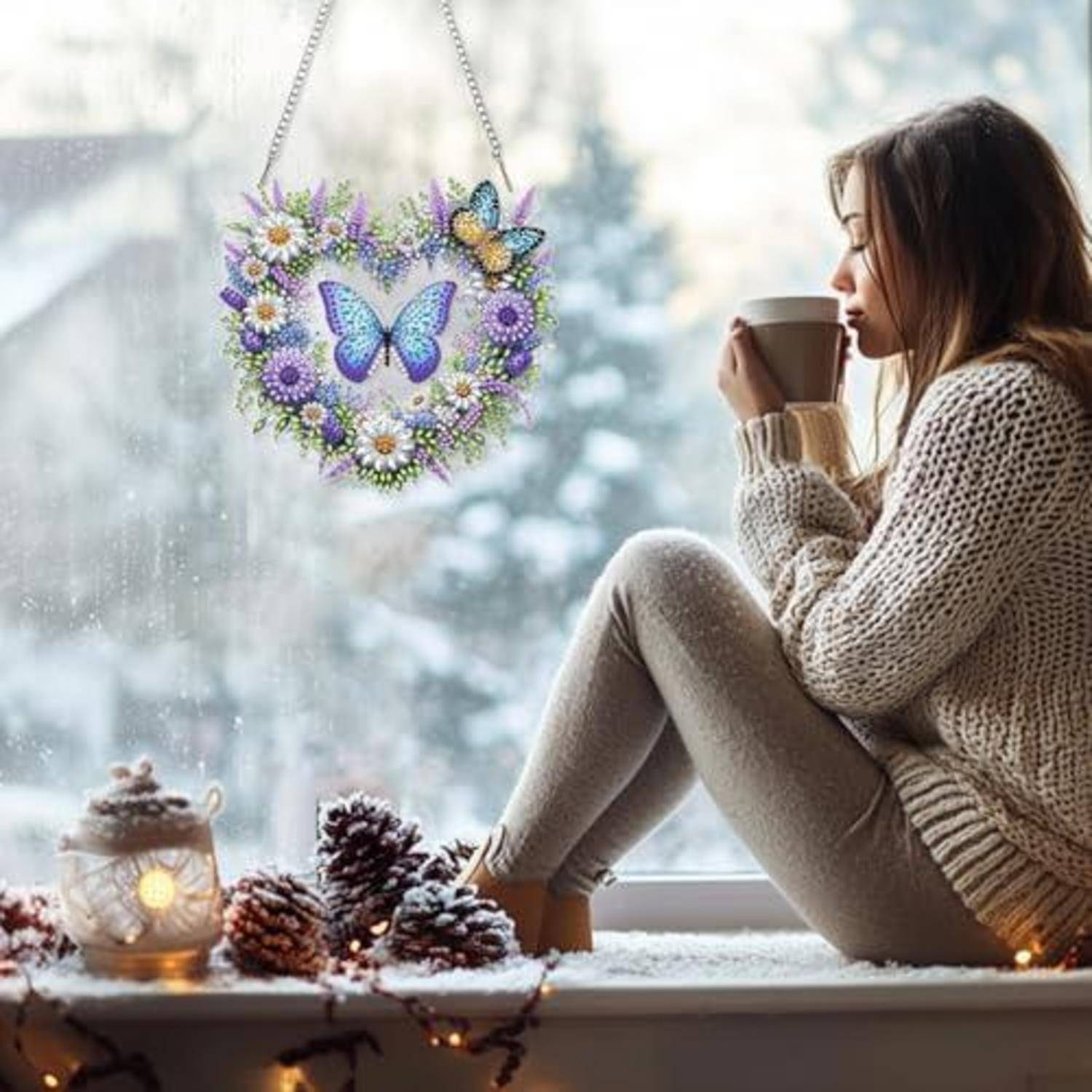 Eine Frau in einem Pullover sitzt an einem verschneiten Fenster, hält eine Tasse in der Hand und schaut auf einen Schmetterlingsreif, der vor dem Glas hängt.