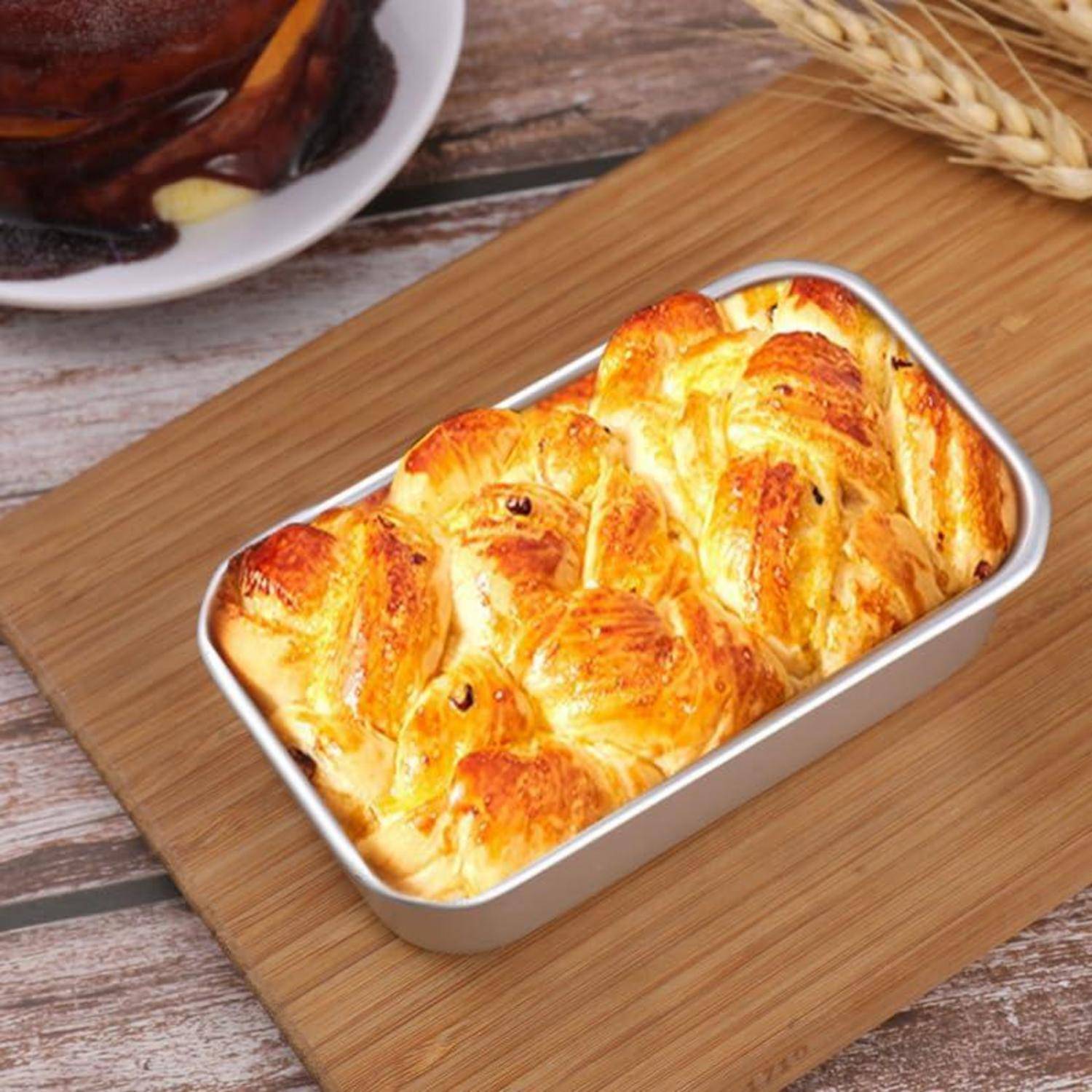 Frisch gebackenes geflochtenes Brot in einer rechteckigen Metallform auf einem Holzschneidebrett, mit einem Glas Tee, das im Hintergrund teilweise sichtbar ist.