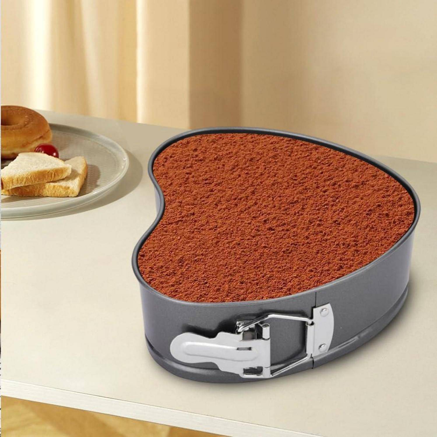 Herzförmige Backform gefüllt mit kakaobestäubtem Dessert auf einem Tisch neben einem Teller mit Bagels und Toast.