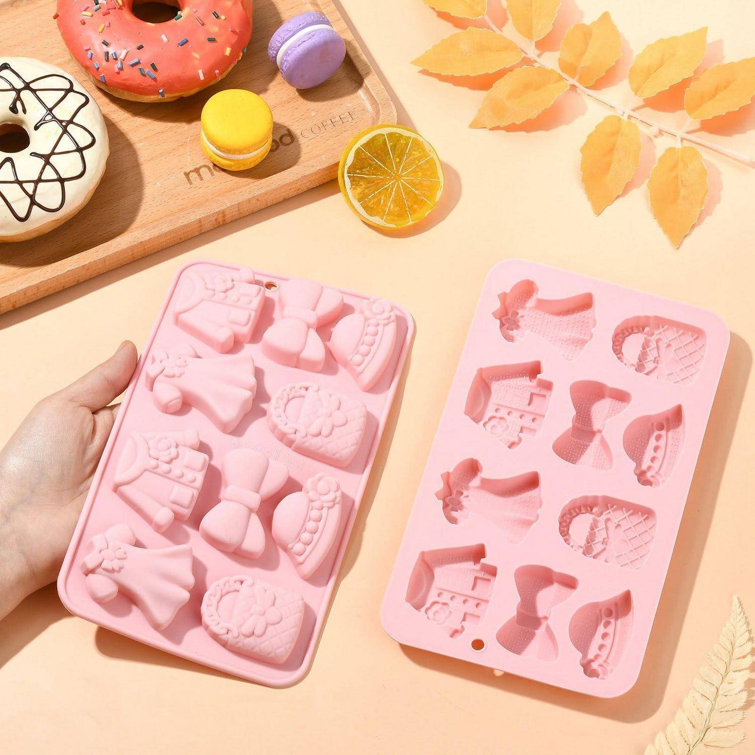 Hand hält rosafarbene Silikonformen mit Schleifer- und Kleidformen neben einem passenden Tablett mit Ausschnitten auf einer pastellfarbenen Tischplatte mit Dessert-Dekorationen.