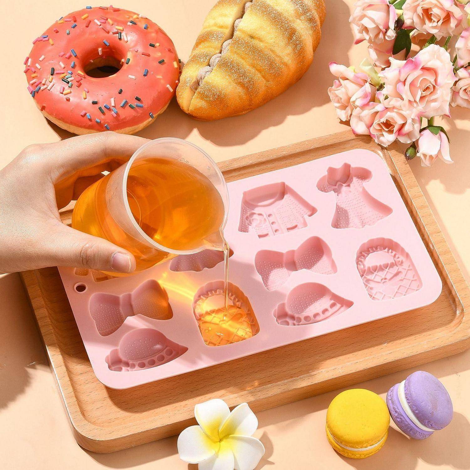 Eine Hand gießt bernsteinfarbene Flüssigkeit in eine rosa Silikon-Gussform auf einem Holzbrett, mit Donut, Croissant, Blumen und Makronen drumherum.