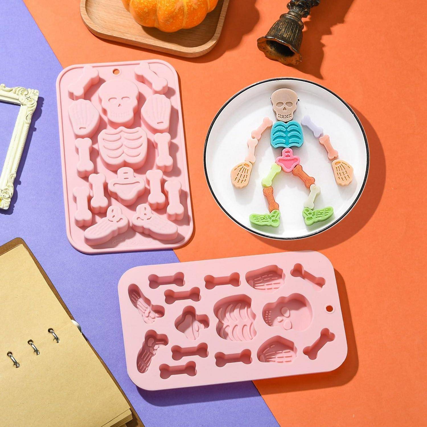 Farbenfrohe Halloween-Backszene mit rosa skelettförmigen Silikonformen, einem Skelettplätzchen auf einem Teller, Kürbissen und einer kleinen Fledermaausdekoration auf orangefarbenem und violettem Papier.