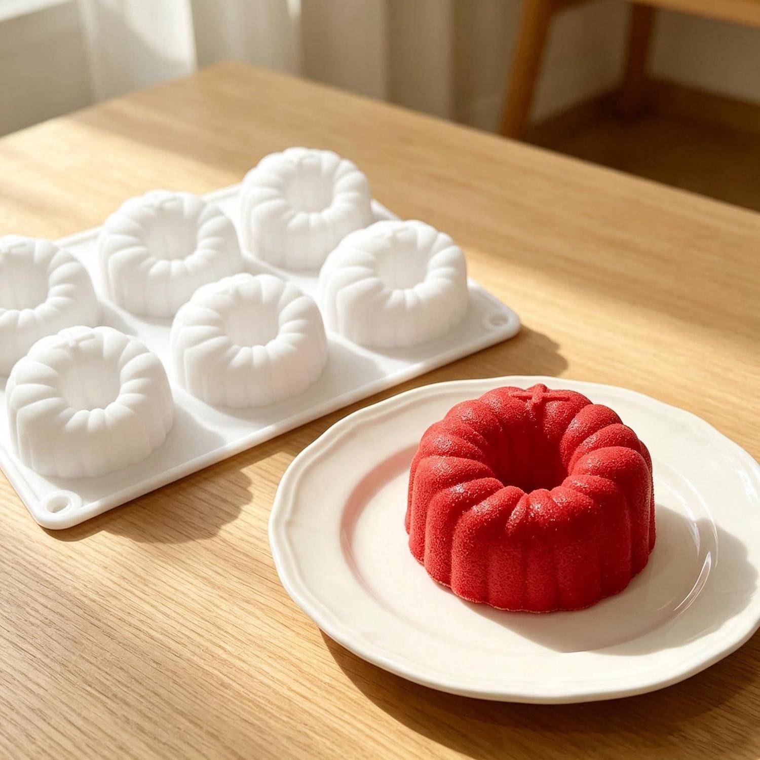 Roter Napfkuchen im Gugelhupf-Stil auf einem weißen Teller neben einem Silikon-Backformtablett mit fünf ausgelösten weißen Kuchen auf einem Holztisch.