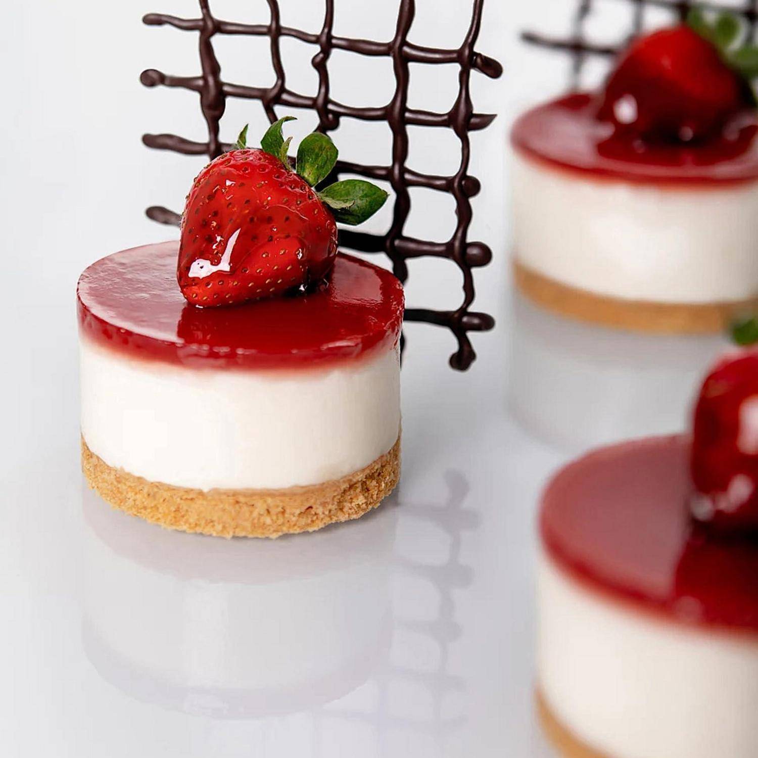 Mini-Erdbeerkäsekuchen mit glänzender roter Glasur, jeder garniert mit einer frischen Erdbeere und einer Schokoladen-Gitterwerk-Dekoration.