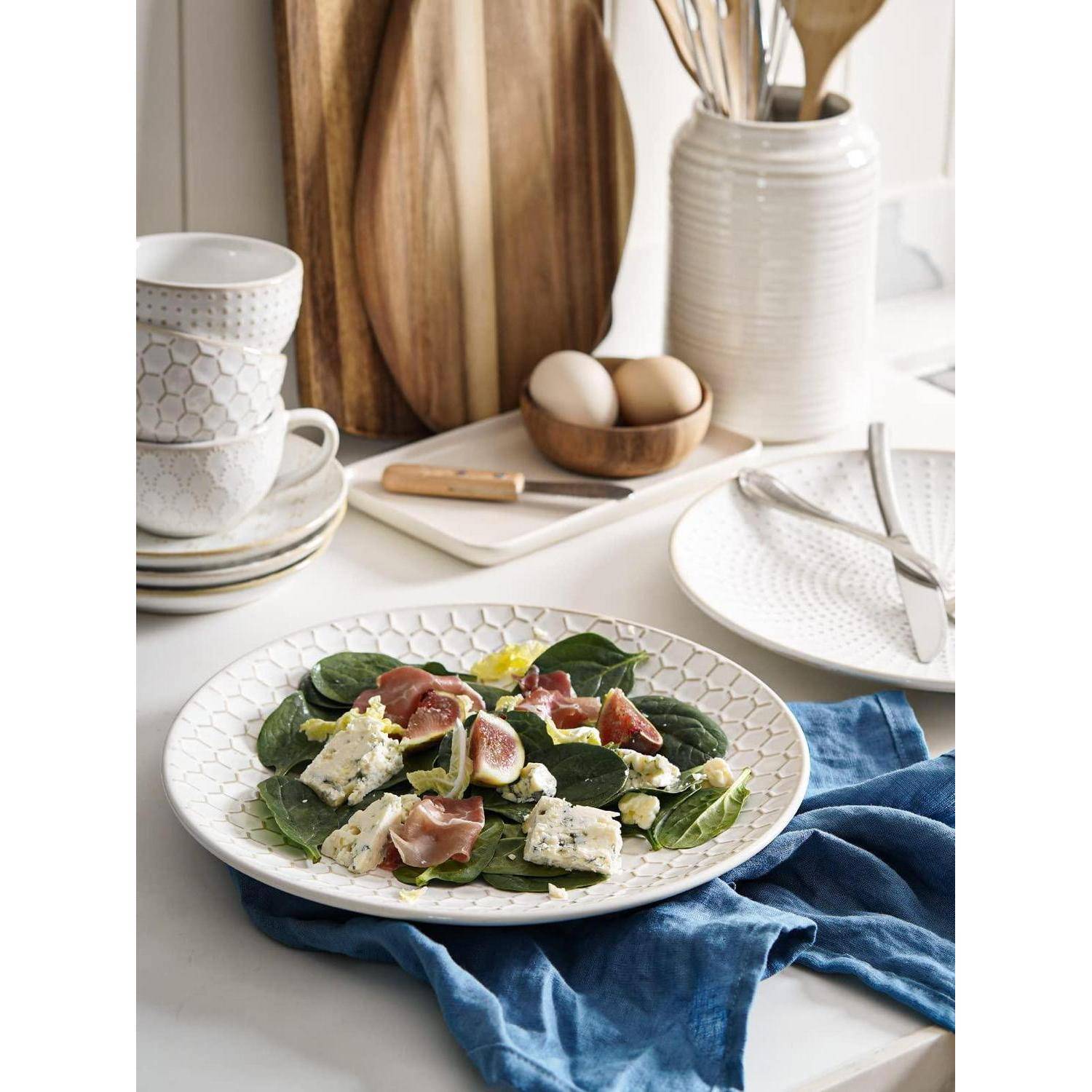 Spinatsalat mit Feigen, Prosciutto und Blauschimmelkäse auf einem weißen Teller, angerichtet auf einem gestylten Küchentisch mit Tassen, Eiern und Besteck.