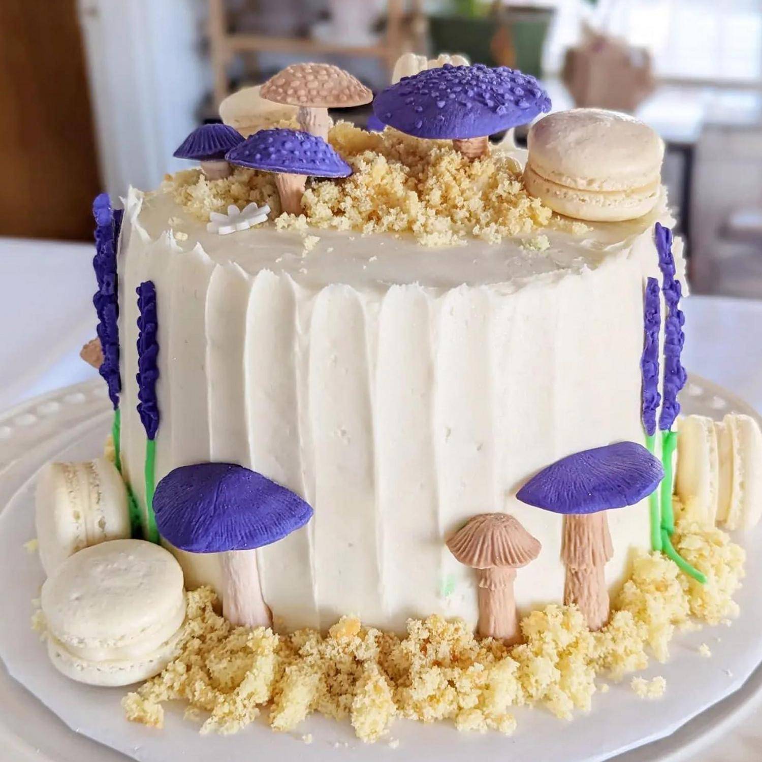 Dekorierter weiß glasierter Kuchen mit violetten Pilz- und Makronen-Toppern auf einem weißen Teller.