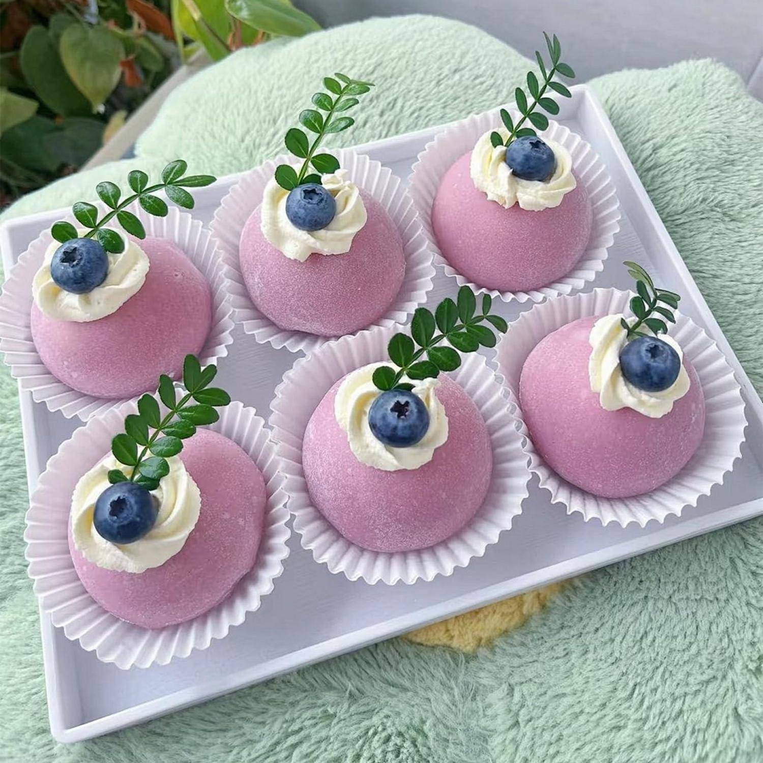Tablett mit sechs rosa Kuppeldesserts mit Sahne, Blaubeeren und grünen Blättern, arrangiert in weißen Papierbechern.