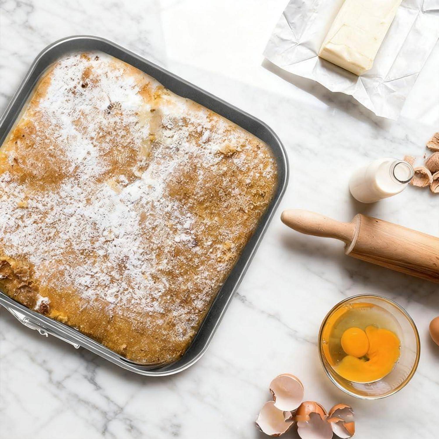 Draufsicht auf einen gebackenen Kuchen oder eine Torte, die mit Puderzucker bestäubt ist, in einer quadratischen Backform, mit Backzutaten und Utensilien in der Nähe.