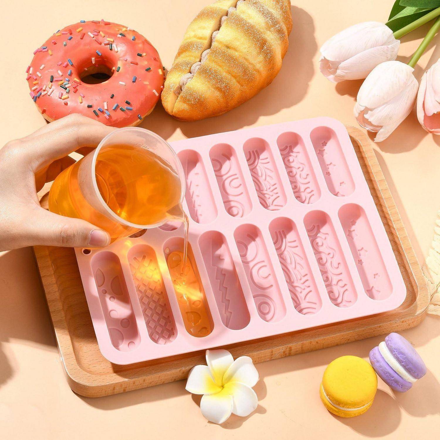 Flüssigkeit, die in eine rosa Silikonform auf einem Holzbrett gegossen wird, umgeben von einem Donut, einem Gebäck, Tulpen und Macarons.