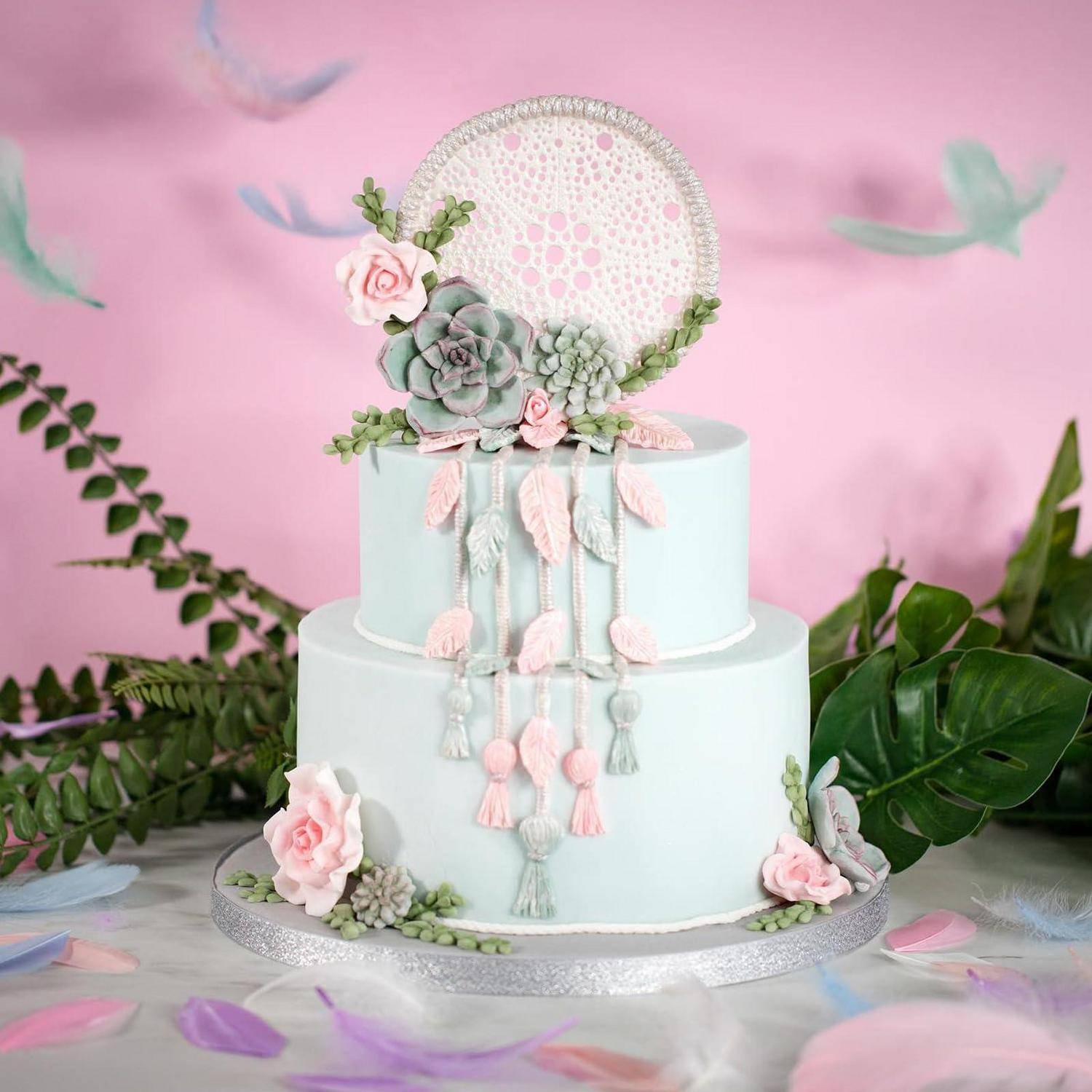Zweistöckiger Pastellkuchen mit Blumen, Sukkulenten, Quasten und einem Spitzenfängerdeko an der Spitze auf einem silbernen Ständer vor einem rosa Hintergrund.