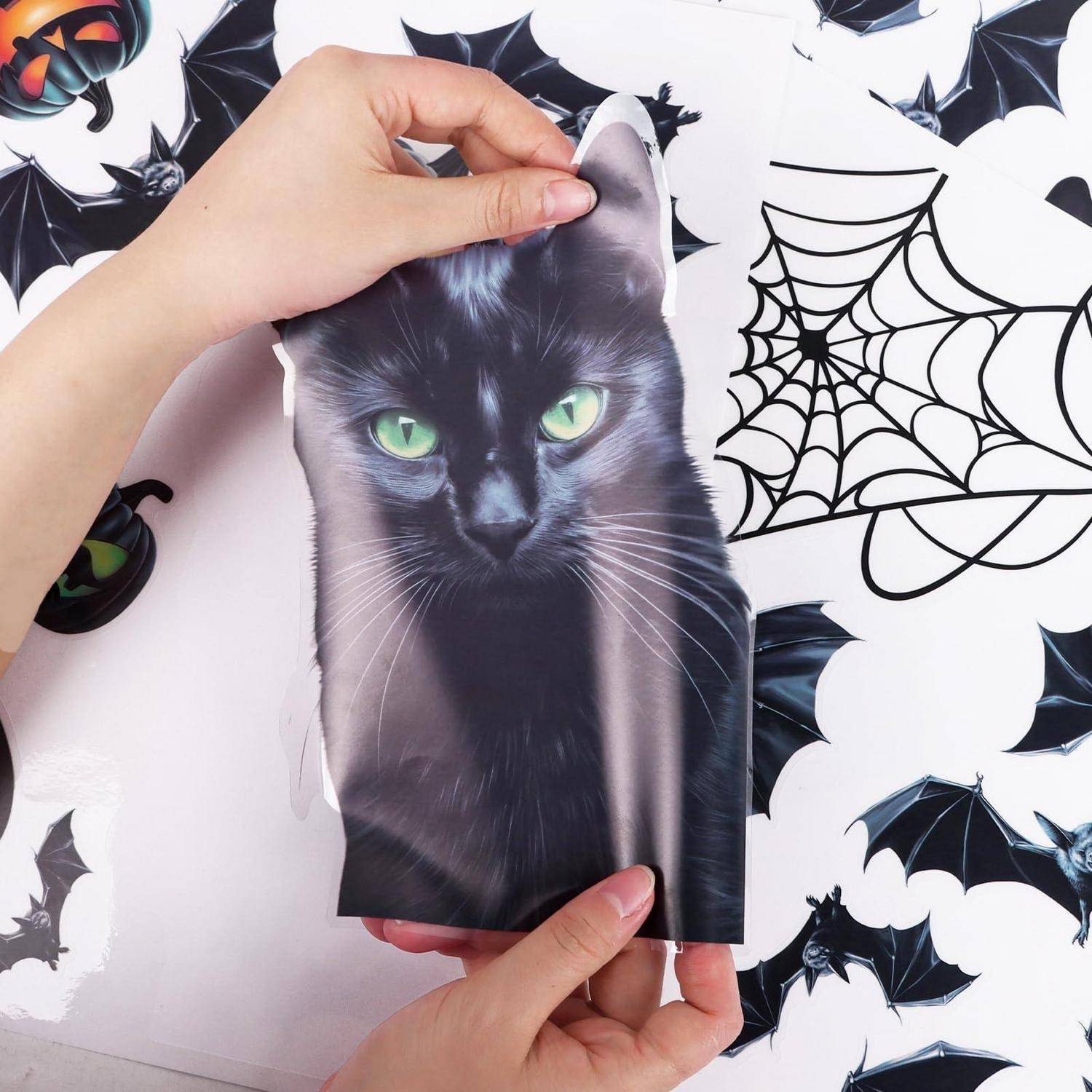 Hände halten ein gedrucktes Bild einer schwarzen Katze über einem Halloween-Hintergrund mit Fledermäusen und einem Spinnennetz.