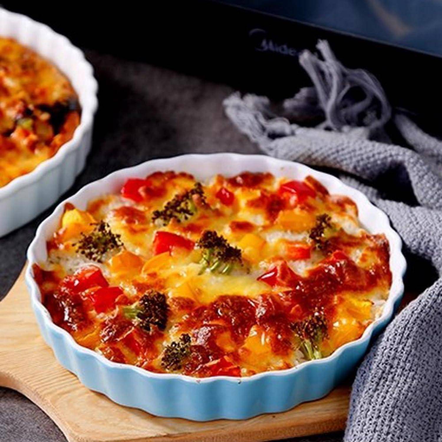 Überbackener Gemüseauflauf in einer blauen Keramikschüssel auf einem Holzbrett, belegt mit geschmolzenem Käse, Tomaten und Brokkoli.