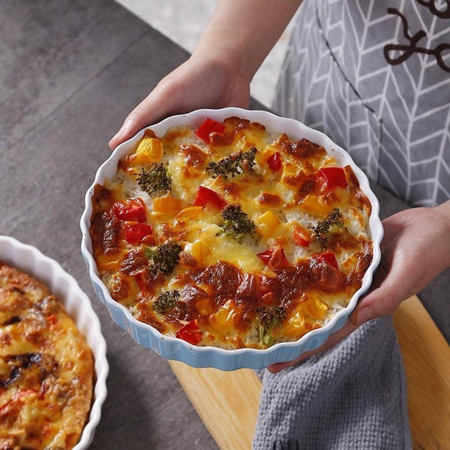 Hände halten eine gebackene Gemüseauflaufform in einer weißen runden Schüssel, bedeckt mit geschmolzenem Käse, Brokkoli und roten Paprika; eine weitere Schüssel ist in der Nähe teilweise sichtbar.