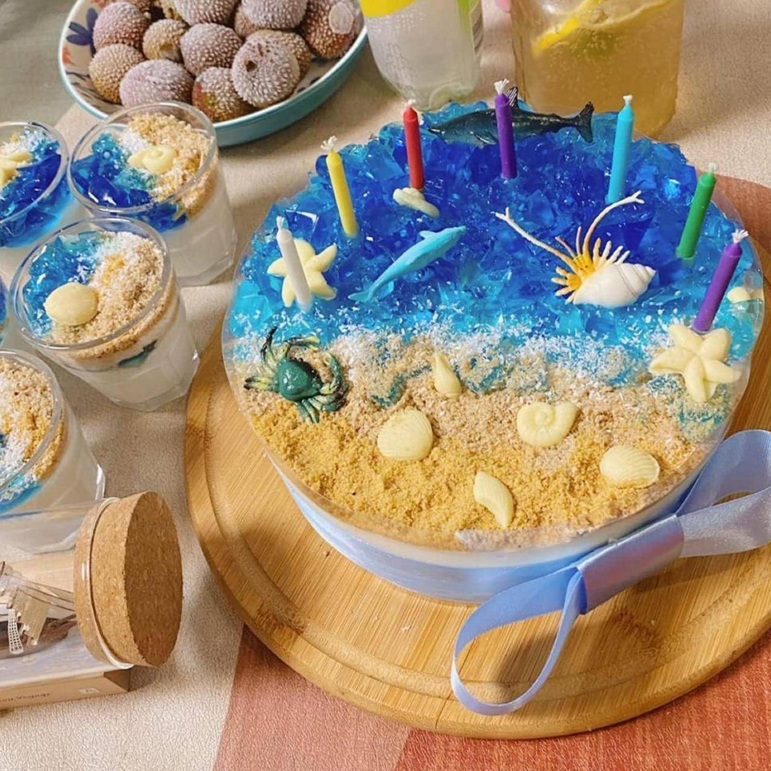 Strandthemen-Kuchen, dekoriert mit blauem Gel, sandähnlichen Krümeln, Muscheln, Seesternen und bunten Kerzen auf einem Holzbrett
