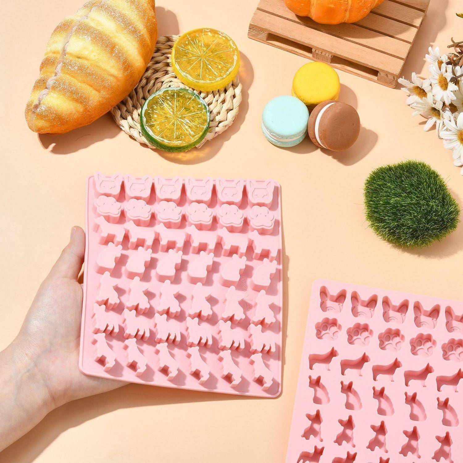 Hand hält ein rosa Silikonform-Tablett mit verschiedenen Tierfiguren, umgeben von Brot, Zitrusscheiben, Macarons und Dekoration auf einer pfirsichfarbenen Tischplatte.